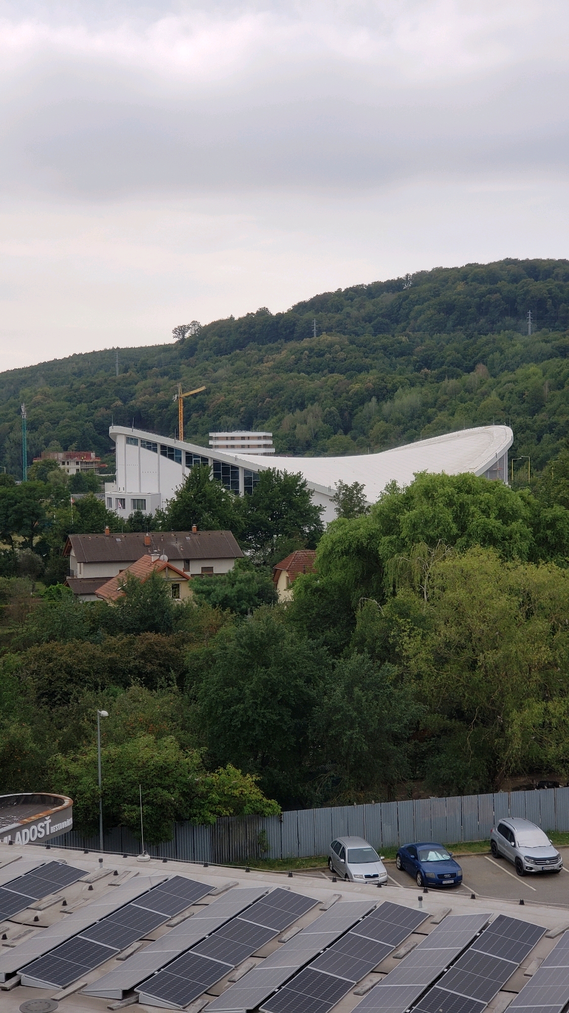 The image depicts a panoramic view of an outdoor urban and natural landscape under an overcast sky. In the foreground, a rooftop installation of numerous solar panels is visible, occupying a significant portion of the lower frame. To the right of the solar panels, a parking area is present with three cars: a silver hatchback, a blue sedan, and a silver SUV. A metal fence runs along the edge of the parking area and extends into the middle ground.
The middle ground is characterized by dense green foliage and trees, interspersed with residential buildings with red-tiled roofs. Beyond the trees, a prominent white building with a distinctive curved roof and large glass windows is visible, suggesting a modern architectural design, possibly a sports or event complex. A yellow construction crane stands near this building, indicating ongoing construction or renovation.
The background consists of a densely forested hill that rises steeply. The trees are predominantly green, indicative of a temperate climate during the growing season. Power lines and poles are sparsely visible on the hillside.
Based on the architectural style of the building with the curved roof and the presence of similar modern structures and infrastructure, this scene is likely located in the vicinity of the Municipal Stadium (Štadión Tatran) in Prešov, Slovakia, which has undergone significant renovations and features a modern, somewhat curved roof structure. The solar panel installation on the rooftop in the foreground is likely part of a commercial or industrial building.