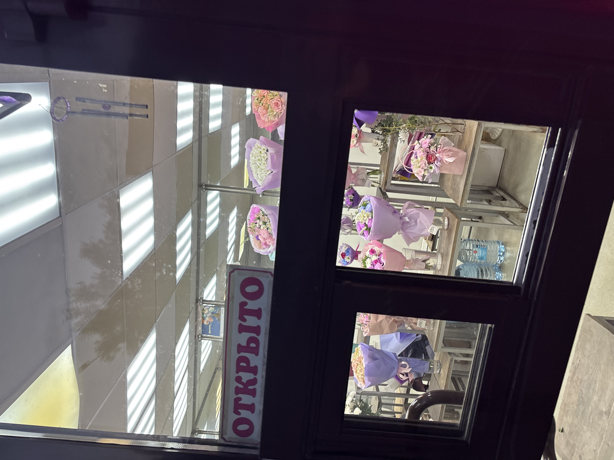 The image shows a view through the window of what appears to be a flower shop. Inside the shop, there are several bouquets of flowers arranged on shelves. The flowers are of various colors and wrapped in paper. There is a sign on the window that says "ОТКРЫТО," which translates to "OPEN" in English. Based on the sign and the presence of flowers, it is likely that this is a flower shop that is currently open. The reflection in the window shows the interior of another building with a geometric patterned wall and ceiling.
Without additional information, the exact location in Saint Petersburg cannot be determined.