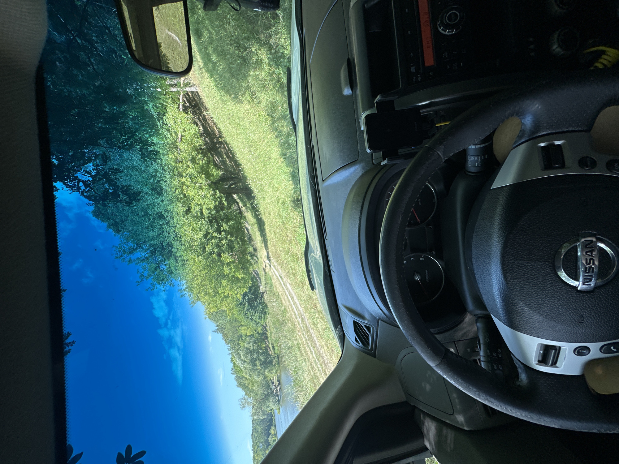 The image is a first-person perspective from inside a Nissan vehicle, looking out through the windshield. The scene depicts a rural or natural environment. Outside the car, a body of water, likely a river or lake, is visible on the left, bordered by a grassy bank and trees. A dirt path or road winds uphill through the grassy terrain on the right, with dense green foliage covering the upper slopes. The sky is a clear, bright blue, with some wispy clouds. The interior of the car shows the steering wheel, dashboard, and a portion of the center console with a car stereo and a phone mount. The overall impression is a drive through a scenic, possibly wooded, natural landscape. The specific location of Makiv, Ukraine, is provided as context for the image.
