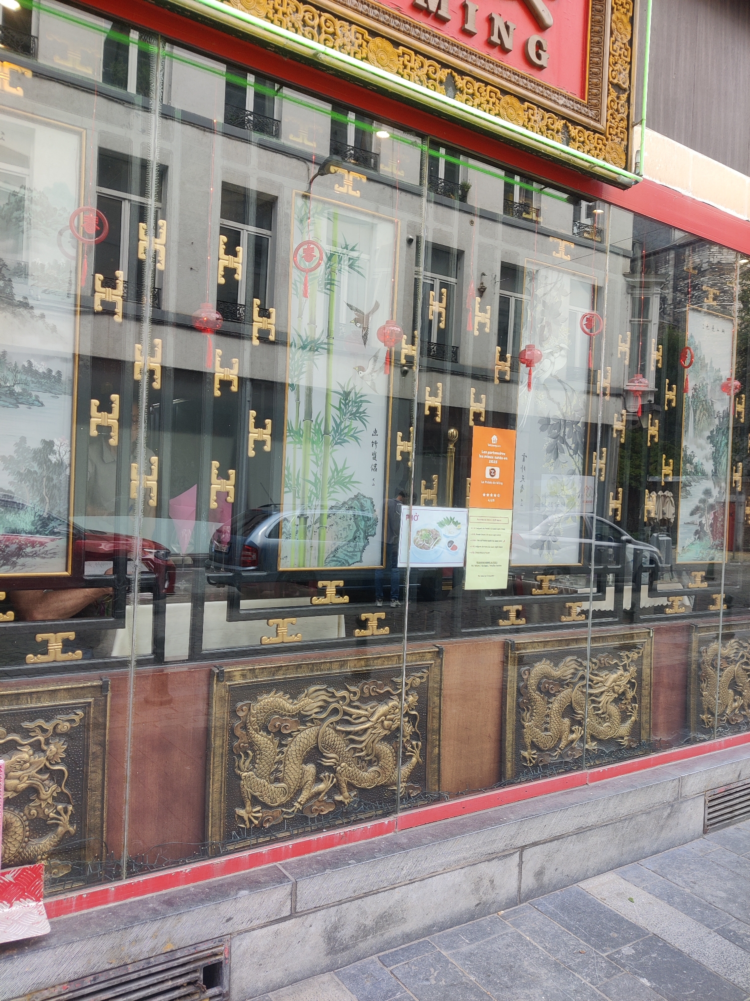 The image shows the exterior of a Chinese restaurant, likely located in Liège, Belgium. The restaurant has large glass windows decorated with ornate gold trim and traditional Chinese artwork, including paintings of landscapes, bamboo, and birds. Red lanterns hang along the top of the window frames. Below the artwork, the window reveals wooden panels with relief carvings of golden dragons. The reflection in the glass shows the facade of a building across the street, with several windows and balconies. Parked cars, one red and one grey, are visible through the lower part of the window, suggesting the restaurant is on a street. A sidewalk made of gray paving stones is in the foreground, with a drain grate visible on the left. The restaurant name, "LE GRAND MING," is visible at the top of the building, outlined in red and gold lettering.