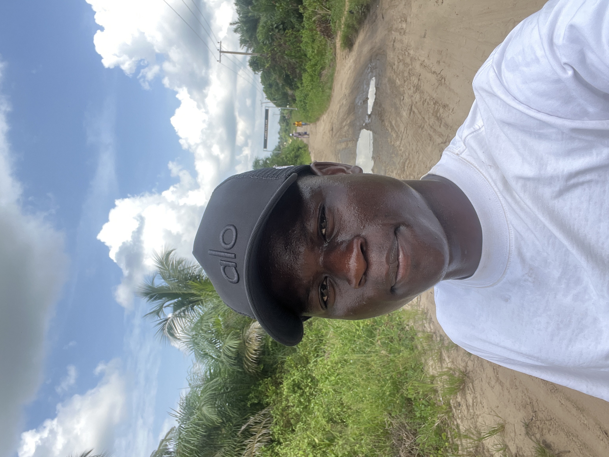 The image shows a man in a white t-shirt and black baseball cap with "ALO" on it, standing on a dirt road. The road has puddles of water and green vegetation on either side. In the distance, other people are walking towards an archway that reads "FARIN VILLAGE." Overhead power lines are also visible. The sky is blue with scattered clouds. The location appears to be a rural area. The city is Tofa, Nigeria.
