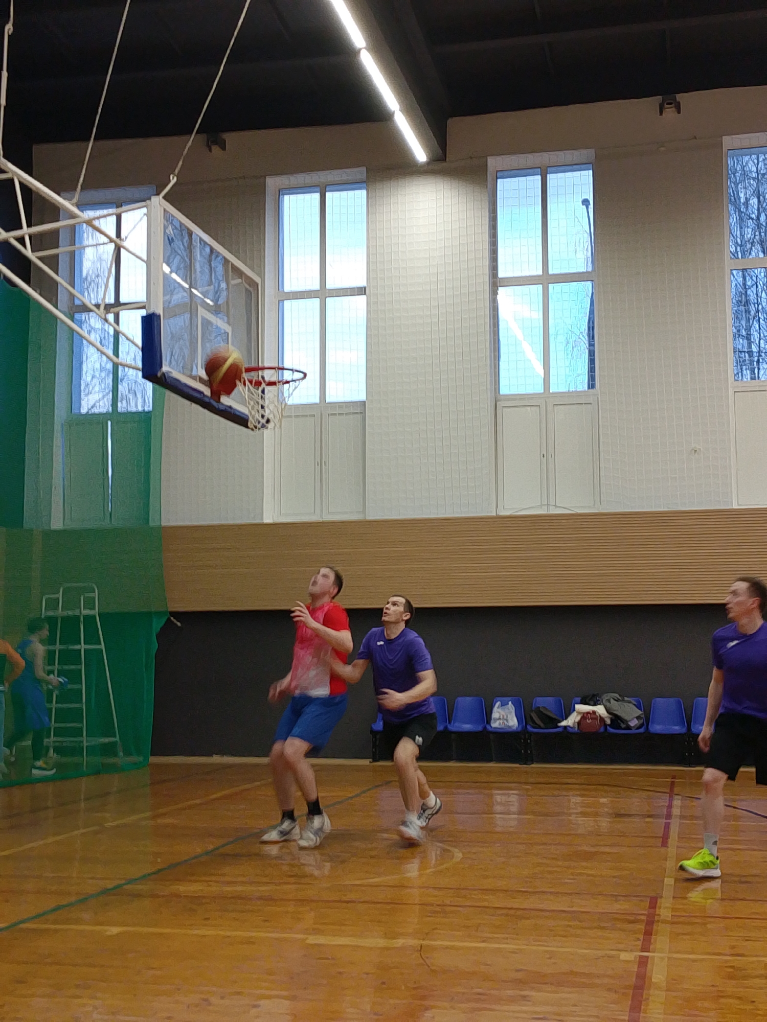 The image shows a basketball game in progress inside a gymnasium. A basketball is entering the hoop. Two men in athletic wear are looking up at the ball near the hoop. A third man in athletic wear is running to the side. Several blue chairs are visible in the background along with a light wood wall. There are several large windows showing trees. The gymnasium appears to be in Izhevsk, Russia.