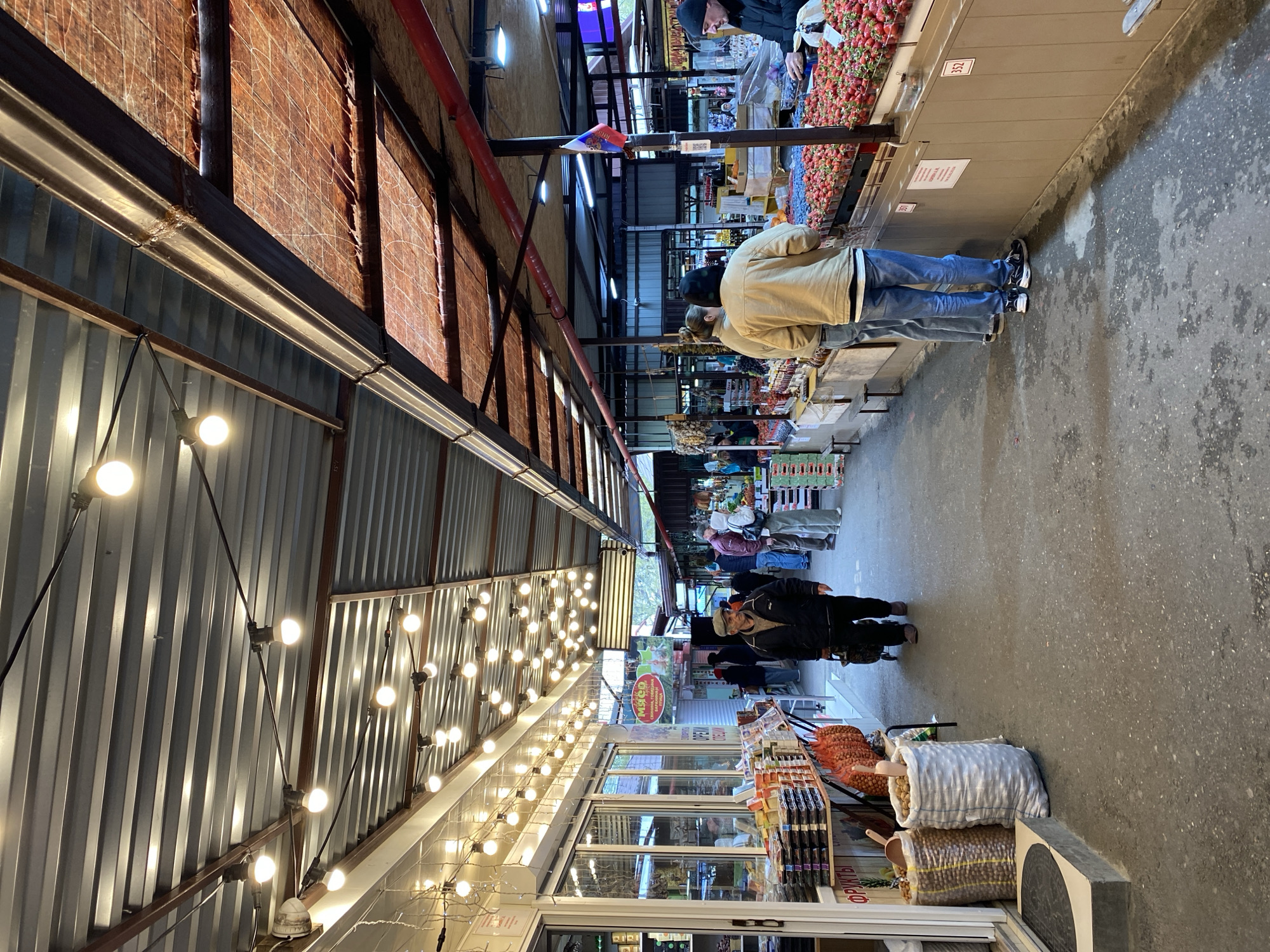 The image depicts an indoor market scene, likely in Gelendzhik, Russia. The market is characterized by a long, narrow aisle with vendor stalls on either side. The ceiling is metal, adorned with strings of incandescent light bulbs. Some stalls are lined with brick, while others are constructed with corrugated metal. Customers are browsing the goods offered, including strawberries, fruits, and vegetables. At the base of the image, bagged produce is displayed for sale. People are present in the aisle, walking and interacting with vendors. The floor is concrete, and the overall lighting appears to be a combination of natural light and artificial illumination from the ceiling fixtures.