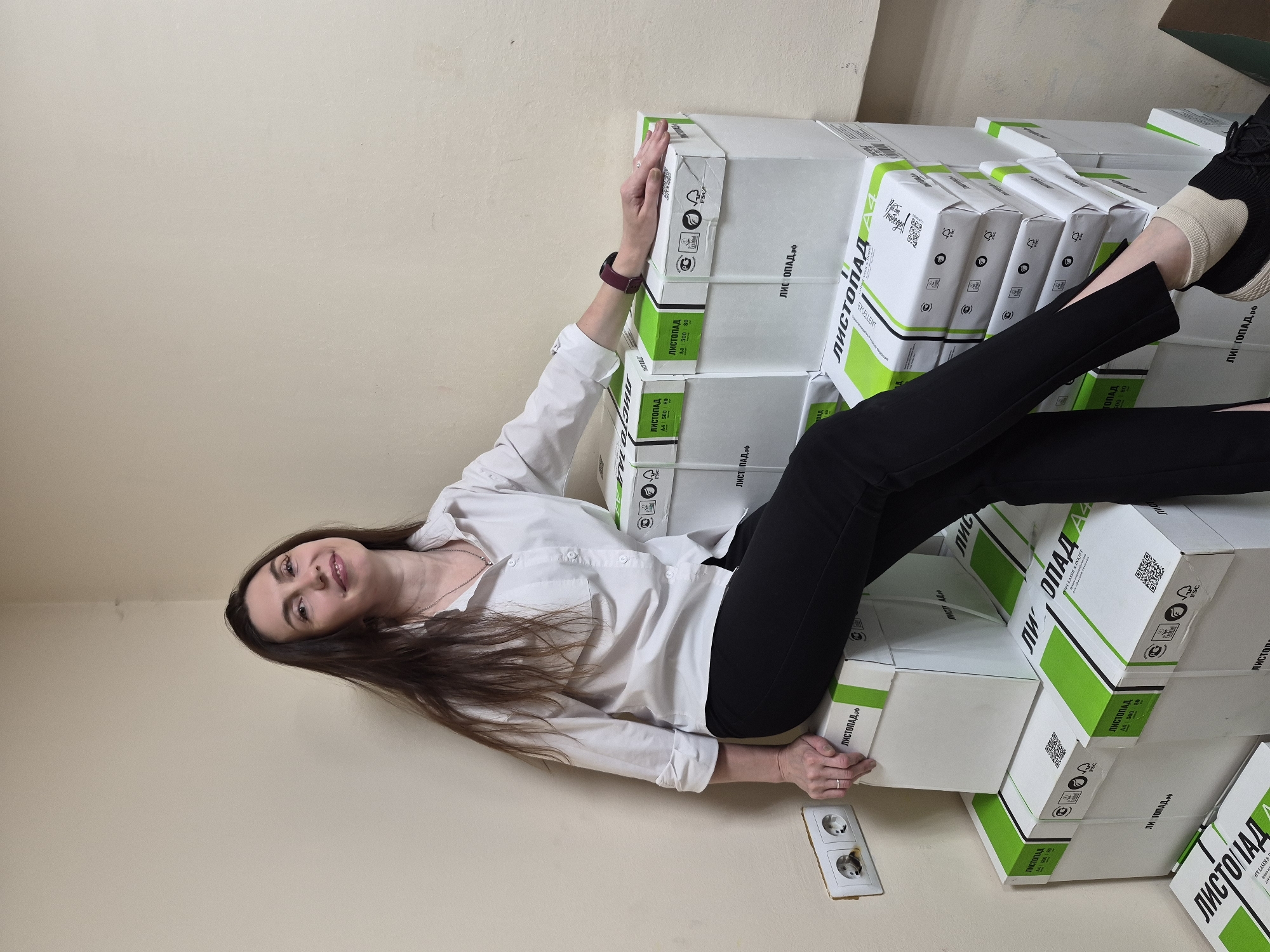 A woman is lying on a stack of white boxes labeled "ЛИСТОПАД" with green accents in an indoor environment with pale beige walls and an electrical outlet. She is wearing a white, button-down shirt and black pants, and her brown hair is loose. She is angled towards the camera with a slight smile. The boxes are stacked along the wall and partially obscure a dark green object in the top right corner.