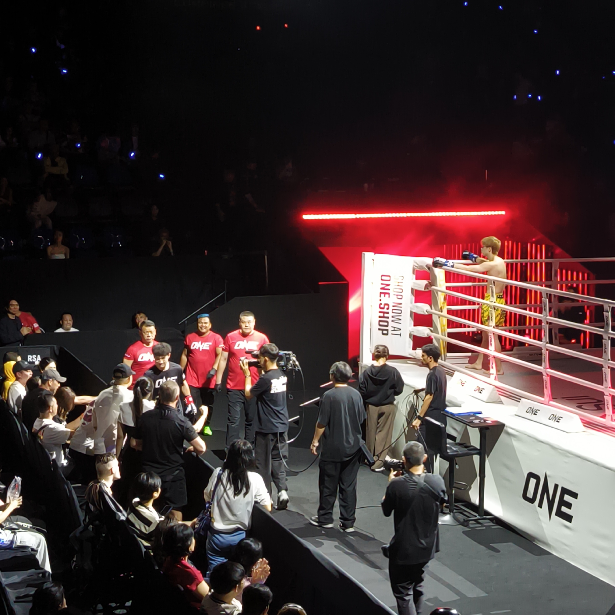 The image shows a boxing ring at an event, likely a One Championship event due to the branding on the ring. The location is presumed to be in Bangkok, Thailand, given the event’s prevalence in that area. A fighter in yellow and black shorts and blue gloves is standing in the ring. To the left, there is a procession of people entering the area, with several wearing "ONE" branded shirts. Several cameramen are filming and photographing the scene. The audience is seated in the background, with some visible in the dimly lit seating area. Above the ring, there is a red light bar.