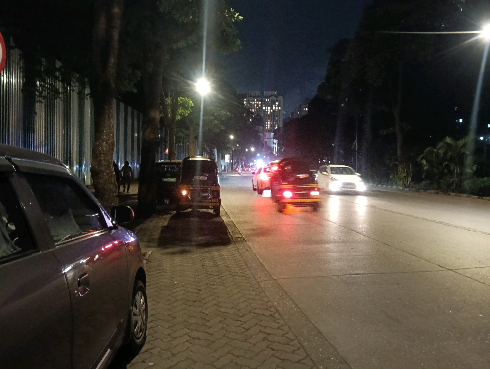 The image depicts a street scene at night in Mumbai, India. A dark-colored sedan is parked on the left side of the frame, its side visible. Beside it, a paved sidewalk is lined with trees and a temporary metal fence. The street itself is wet, reflecting the streetlights and the taillights of moving vehicles. Several cars are visible on the road, including a light-colored sedan and a darker vehicle with its brake lights illuminated. In the background, a multi-story building with many illuminated windows is visible. The overall atmosphere is one of an urban environment at night, with traffic and city lights.