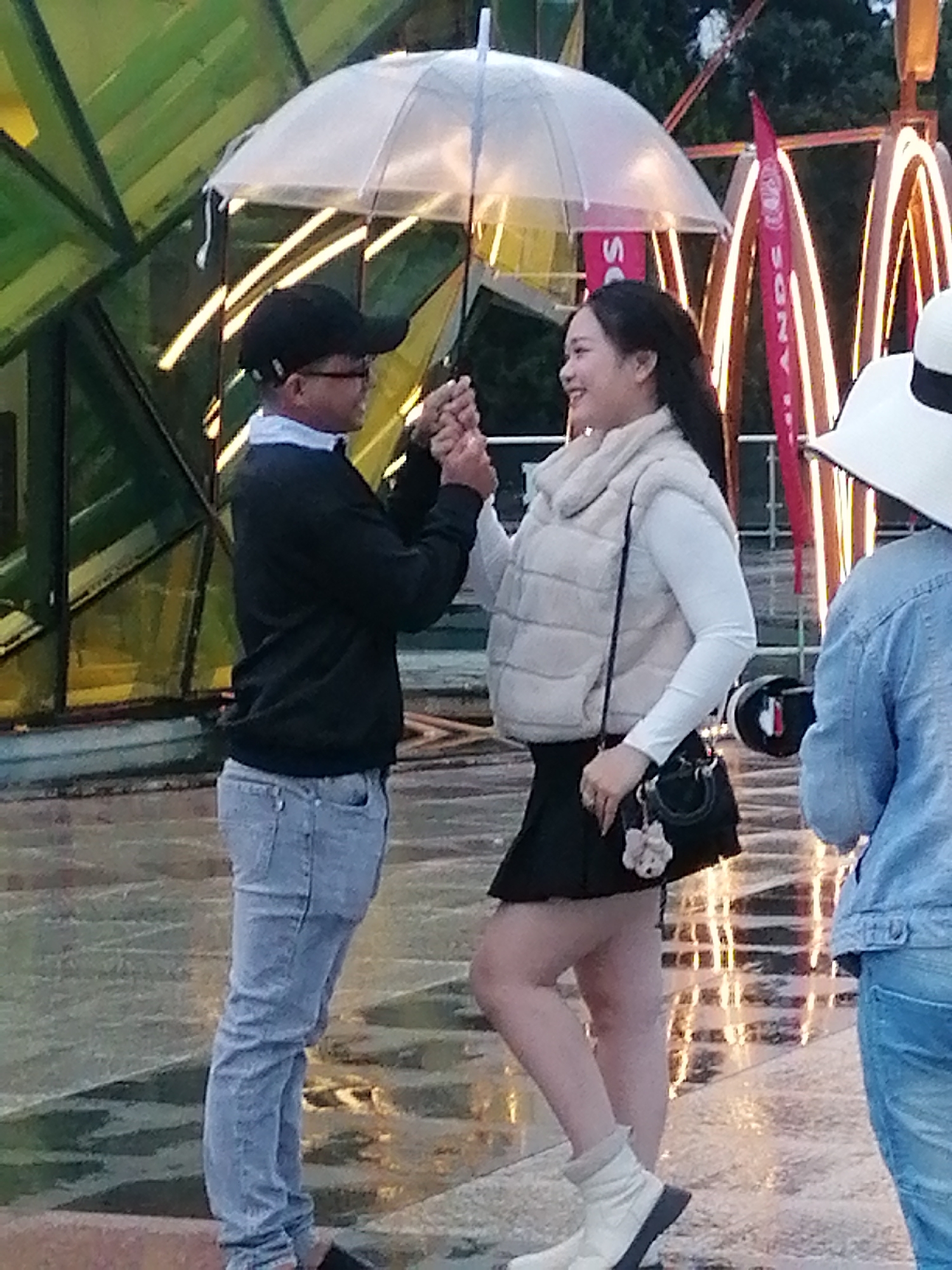 In Da Lat, Vietnam, a couple stands under a transparent umbrella, holding hands. The man, wearing a black cap, glasses, a dark sweater over a light collared shirt, and light blue jeans, faces left. The woman, in a beige puffer vest over a white long-sleeved shirt and a black skirt, with white boots, faces right. Behind them, a bright, abstract green structure is visible, along with illuminated decorative arches that suggest a nighttime or dusk setting, possibly a public gathering space or entertainment area. The ground appears wet, indicating recent rain.