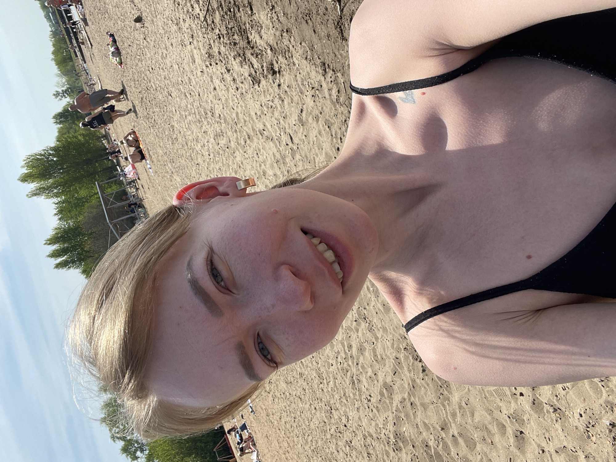 A blonde woman is pictured in a close-up selfie at a beach. She is fair-skinned, with light blonde hair, wearing a black swimsuit. In the background, there is a sandy beach with people in various activities such as laying in the sun, walking around, and standing by the structures. The sky is partially visible with a few clouds. Trees are seen along the edge of the beach. The location appears to be a beach in the city of Perm, Russia.