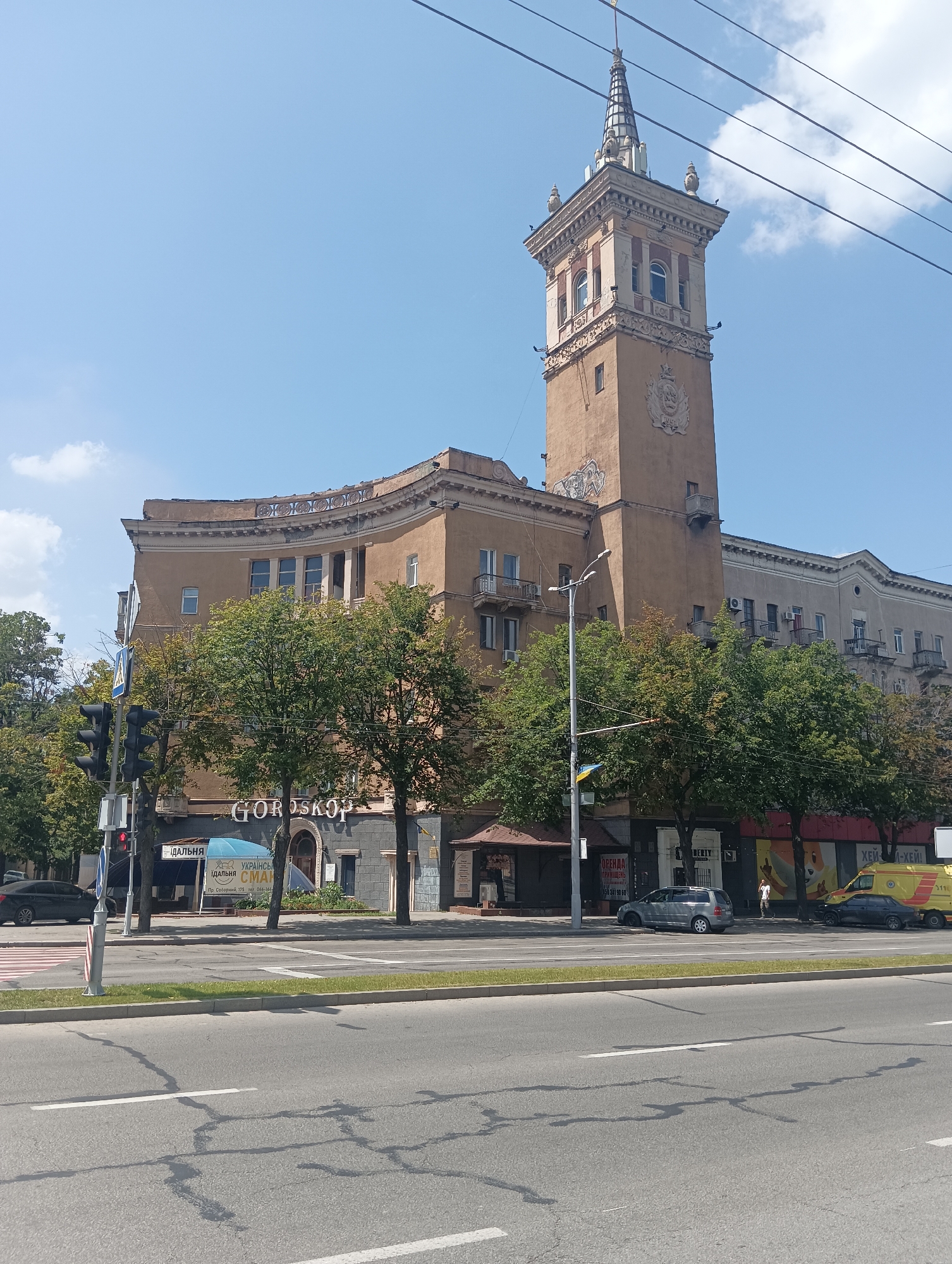 A wide, low-angle, exterior, daytime shot captures a portion of a city block featuring a distinct tower and a curved building. The tower, a prominent feature, is tall and segmented, with a conical spire topped by a finial. The tower's facade is a muted beige and features decorative elements, including a crest and a recessed balcony. Adjacent to the tower is a curved, light brown building with multiple stories, featuring balconies and windows. The lower levels of the building house shops, with visible signage including "GOROSKOP" and other Cyrillic text. Green trees with full canopies are positioned in front of the buildings. A traffic light with three red lights visible stands at an intersection to the left of the frame. A crosswalk with white stripes is present in the foreground, spanning the width of the road. The road itself is asphalt with white lane markings, and shadows of overhead wires are cast upon it. Several cars are parked or driving on the road, including a black sedan, a silver SUV, and a yellow service vehicle. The sky above is a clear blue with a few scattered white clouds.