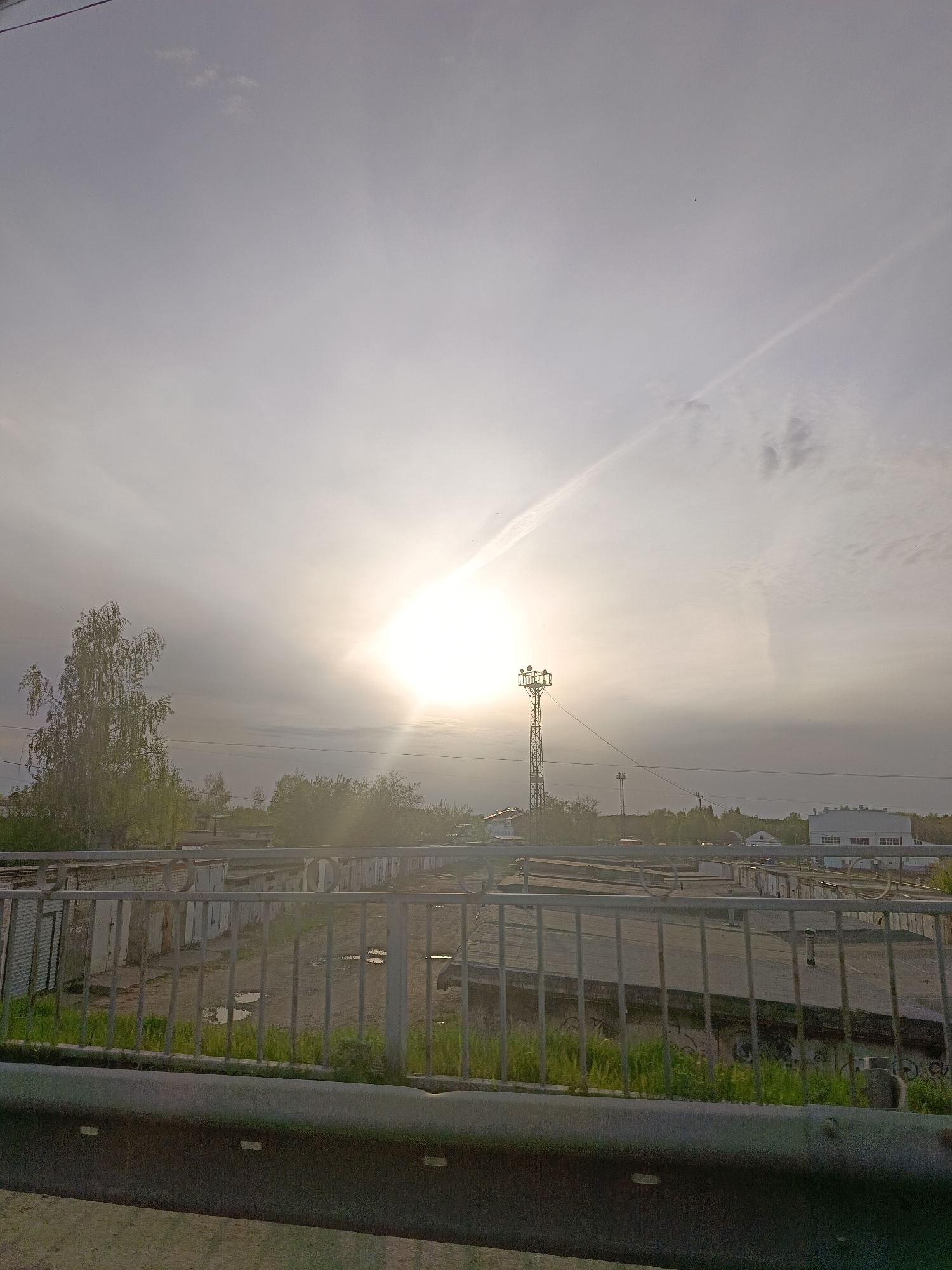 The image presents an overcast day over a garage complex in what is likely Chaikovsky, Russia, based on visual cues. The sun is obscured by a layer of clouds, yet still bright, slightly left of the frame's center. A metal lattice communication tower stands in the midground, further back than the rows of garages. A gray metal railing with green grass below it is in the foreground. The view is from a slightly elevated position, possibly a bridge. The garages appear to be concrete with metal doors. The sky is a uniform gray with a possible contrail extending toward the upper right corner. Several buildings, probably low-rise residential or industrial buildings, appear behind the garages and trees in the distance. There is no evidence of people or activity in the immediate scene.