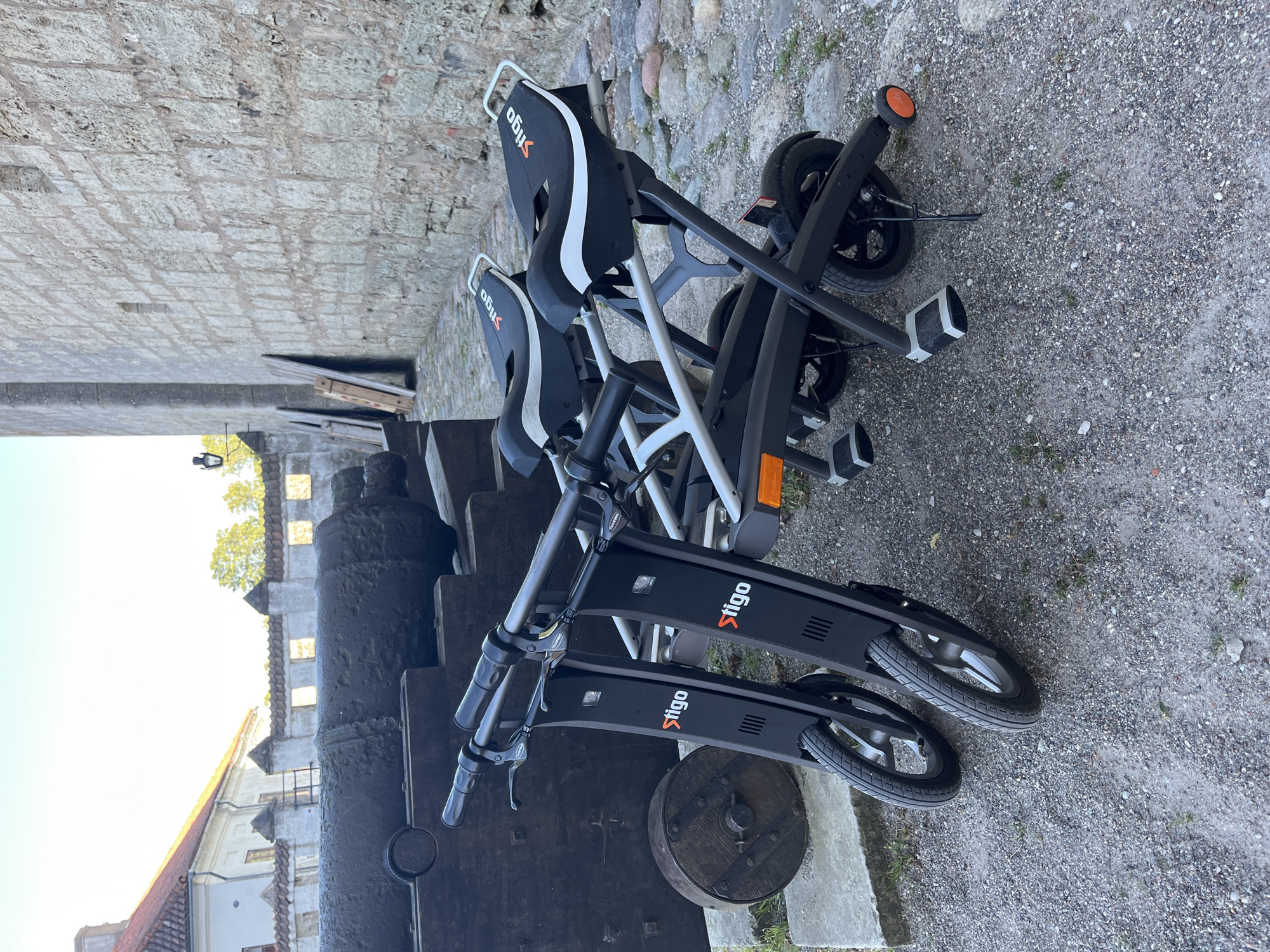 The image depicts two black electric scooters parked next to a large, old cannon on a cobblestone ground. The scooters have "Stigo" branding on them. In the background, there is a stone wall characteristic of medieval architecture, and a white building with a red-tiled roof. The setting suggests a historical or tourist location. The scene is an outdoor shot under natural light, likely during the daytime. The scooters are positioned in the foreground, while the cannon and the stone wall occupy the mid-ground and background, respectively. The location appears to be Kuressaare Castle in Kuressaare, Estonia, a well-known landmark with historical cannons and architecture. There are no people visible in the image.