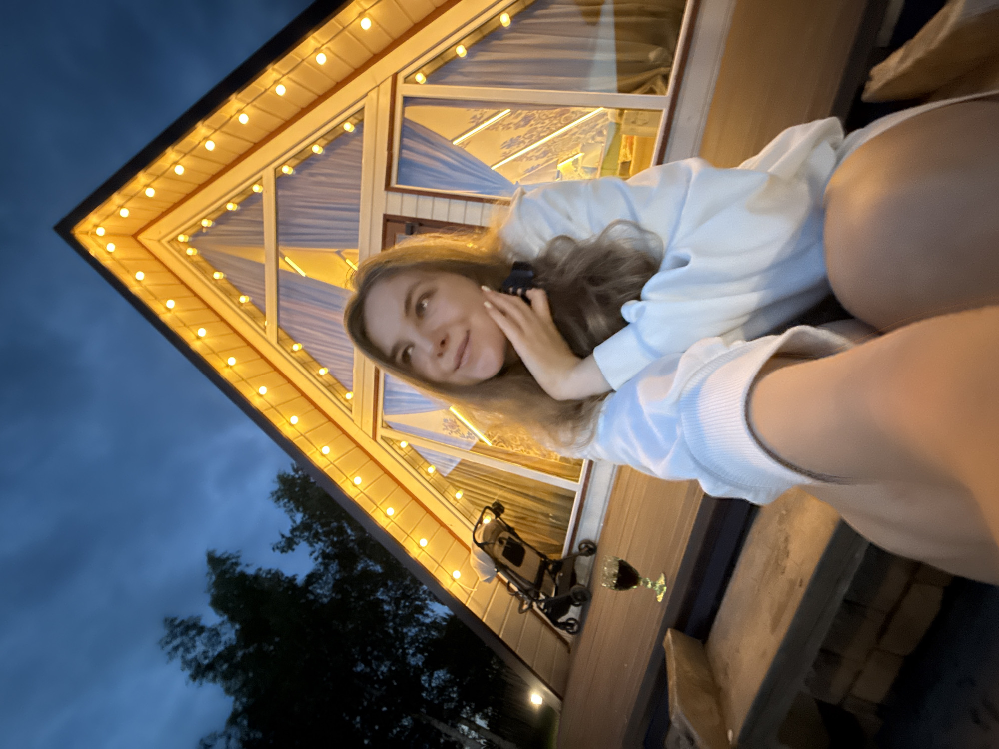 The image, captured in Suzdal, Russia, depicts a young woman posing for a selfie outdoors during the evening. The woman, with long, light brown hair, is wearing a white sweatshirt. She smiles softly at the camera, resting her chin on her hand, with a relaxed and pleasant expression. The perspective is a close-up selfie, with the photographer's hand and part of a leg visible in the immediate foreground.
Behind her is a distinctive triangular-shaped building, possibly an A-frame cabin or house, illuminated by warm string lights running along its roofline. Through the large windows of the building, curtains are visible inside. The dark blue sky of dusk or early night forms the background, with the silhouettes of trees visible towards the lower left.
On a wooden surface, likely a balcony or deck, in front of the building, a wine glass containing a dark liquid and a baby stroller or pram are present. The overall scene suggests a cozy and relaxed evening atmosphere in a natural setting. There is no visible text in the image.