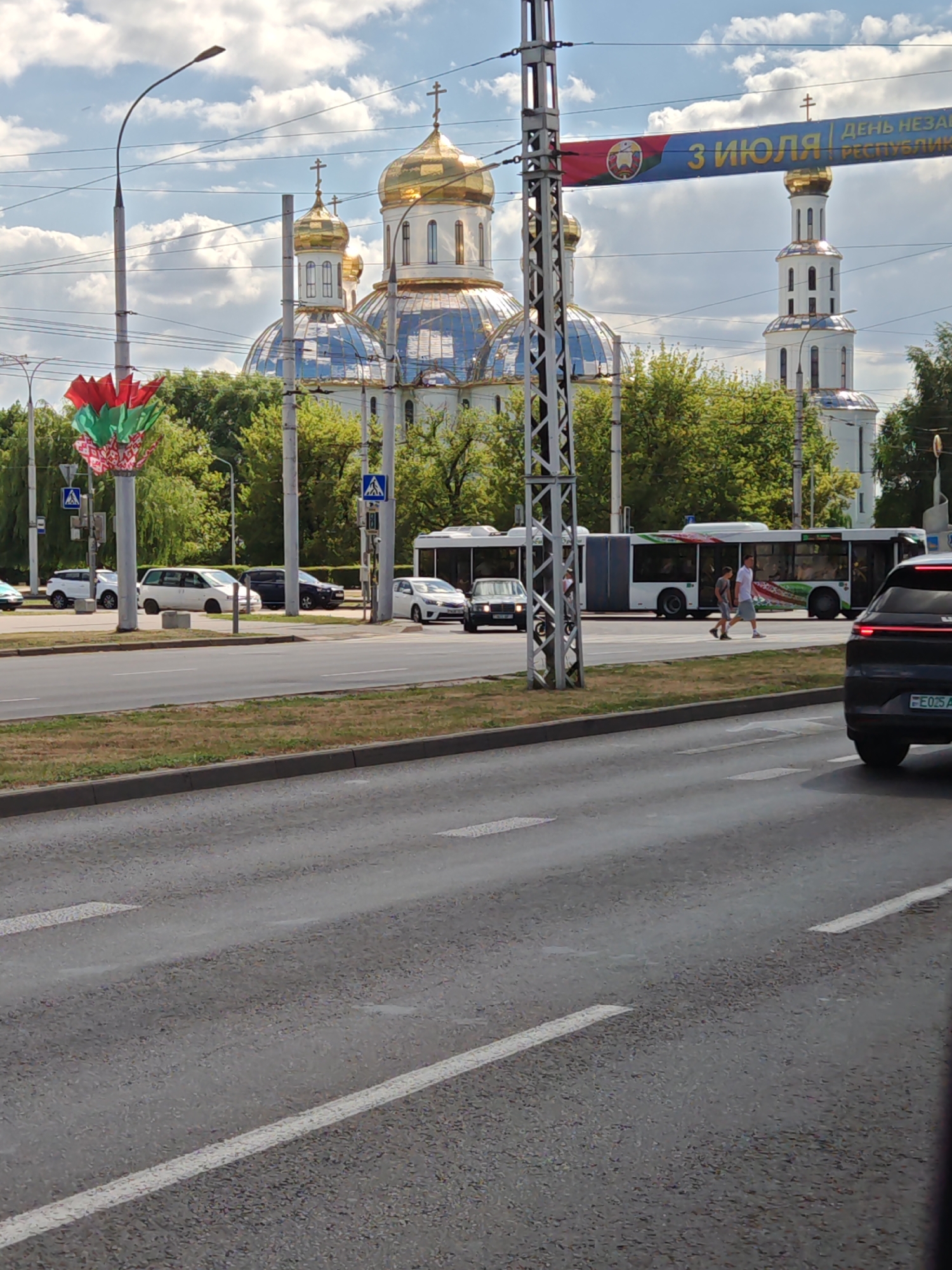 In the city of Alexandrovka, Russia, on a clear day with scattered clouds, a street scene unfolds. Dominating the background is a large, white Orthodox cathedral with multiple golden domes, reflecting the sky. To the right of the cathedral stands a tall, white bell tower, also topped with a golden dome. A banner with Cyrillic text and the Belarusian flag is suspended overhead, likely indicating a holiday or event.
In the mid-ground, a modern, articulated bus with a colorful design on its side is stopped, with passengers visible inside. Two individuals are walking across a pedestrian crossing in front of the bus. Several cars are parked and driving on the road, including a black SUV in the foreground. A street lamp with a decorative red, green, and white element is visible on the left. The environment includes a paved road, a grassy median, and a sidewalk lined with trees. The overall impression is of a bustling urban environment with significant cultural and religious landmarks.