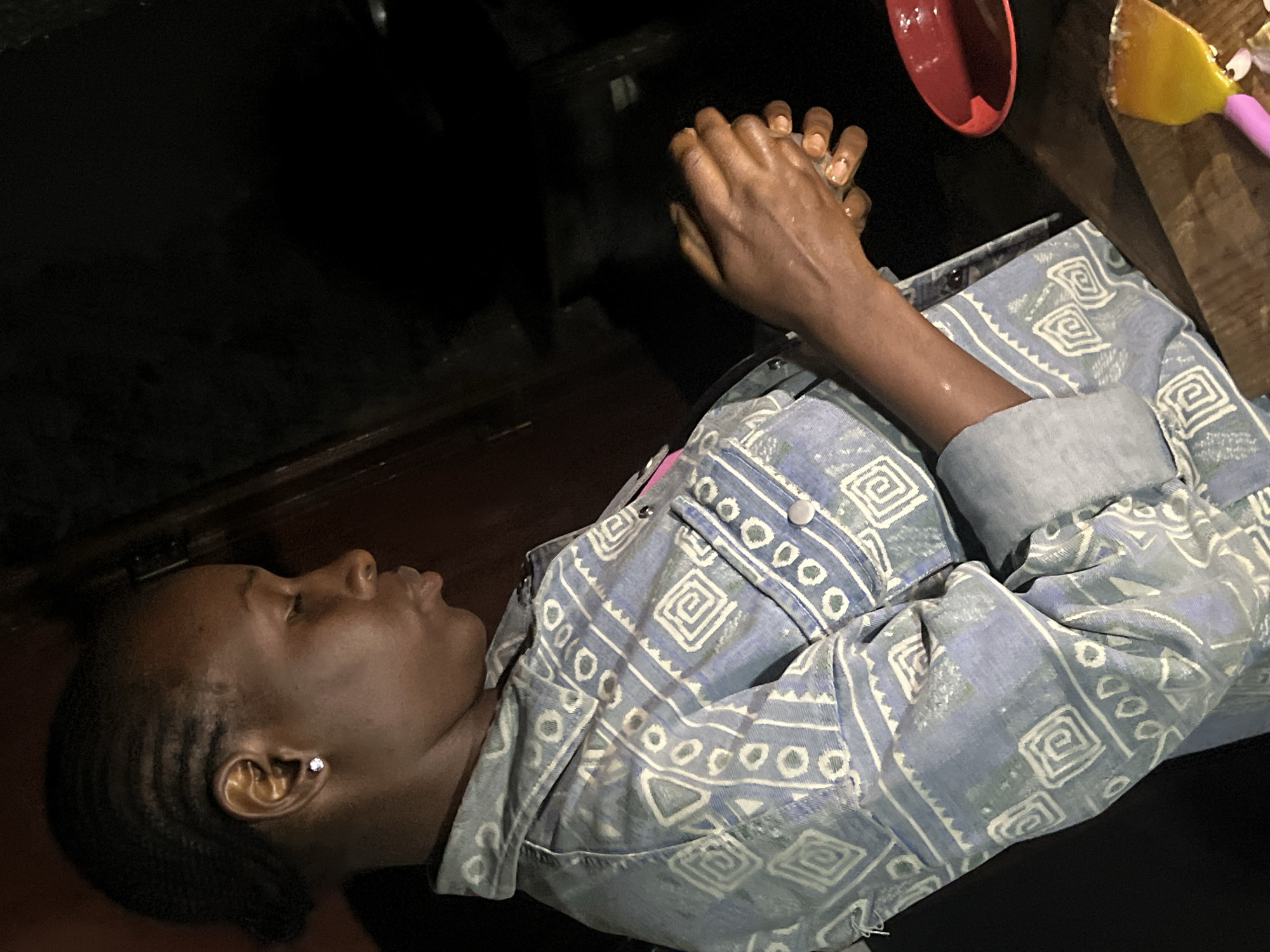 A person is seen in the foreground, facing right in profile. They have dark skin, short braided hair, and are wearing a light blue denim jacket with a geometric pattern. The person is holding their hands in front of their chest, appearing to be working with them. The background is dark and out of focus, with some hints of a wooden surface and a red bowl in the lower right corner. The lighting suggests an indoor setting, possibly at night. The image does not provide any information about the specific location in Durumi, Nigeria.