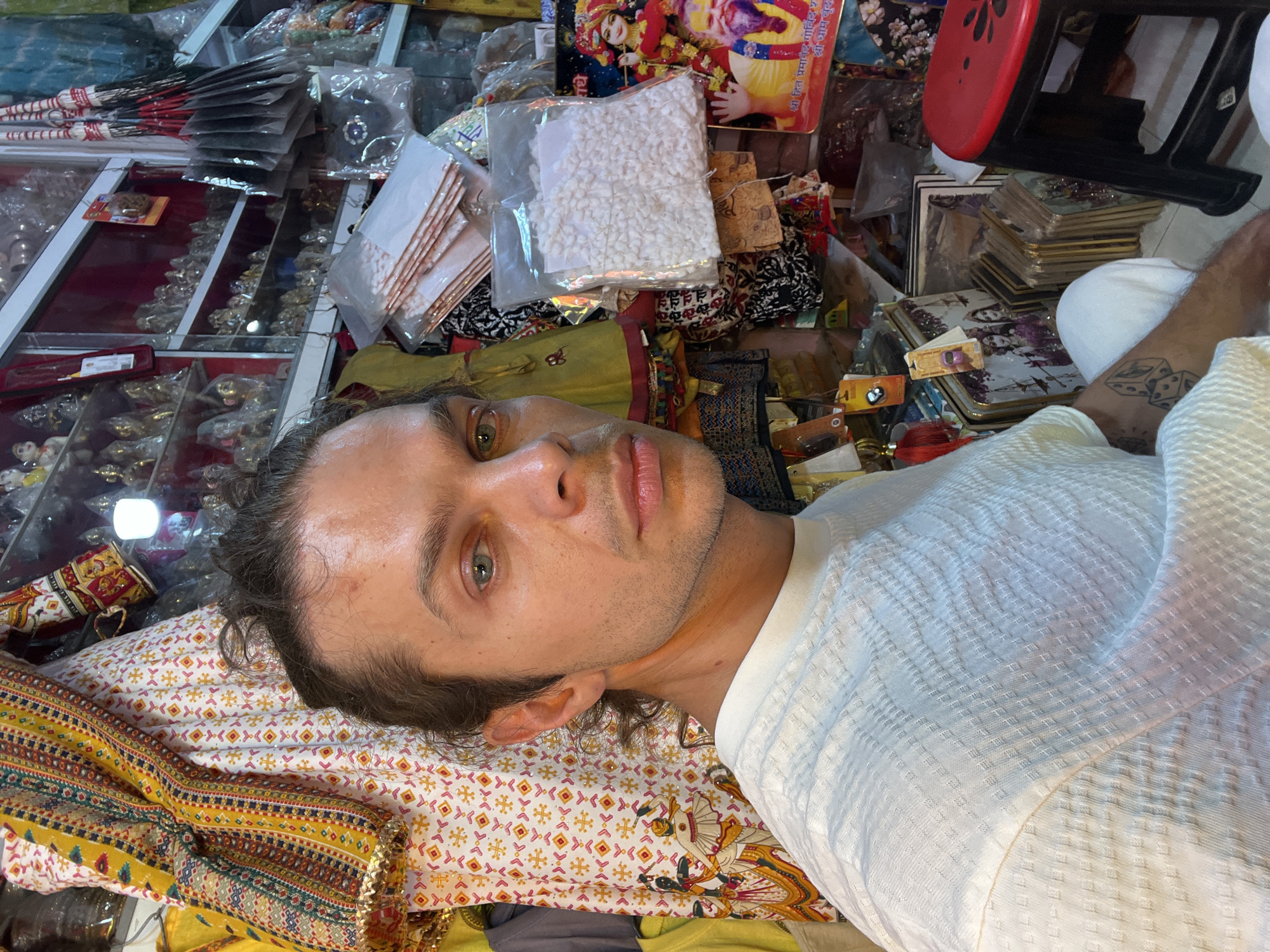 The image shows a man lying down amidst a cluttered environment, possibly within a shop in Mathura, India. The man has fair skin, light-colored eyes, and is wearing a white, textured shirt. He has a tattoo on his arm. He is lying on a patterned cloth, with various items scattered around him, including religious paraphernalia, packaged goods, and what appears to be a display case filled with small items. A red stool is visible in the background, along with other objects like umbrellas and a stack of cards or books. The setting is characteristic of a busy, commercial area with a religious focus.