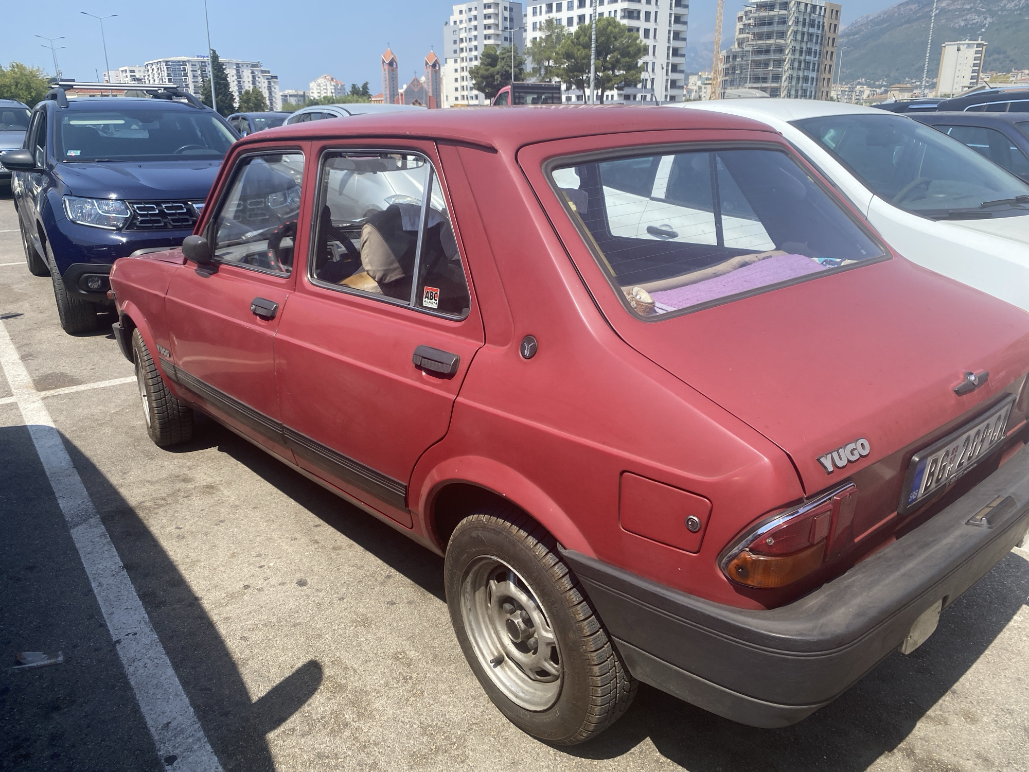 The image shows a red Yugo car parked in a lot in Bjeliši, Montenegro. The Yugo is positioned diagonally, with its rear facing the viewer. A dark blue SUV is parked to the left of the Yugo. In the background, several tall buildings and construction cranes are visible, indicating an urban environment. The ground is paved with asphalt, and parking lines are present. The sky is clear and blue, suggesting a sunny day. The Yugo has a visible license plate, and a purple blanket is draped over its rear window. A sticker with "ABC ALARM" is on the rear passenger side window of the Yugo.
