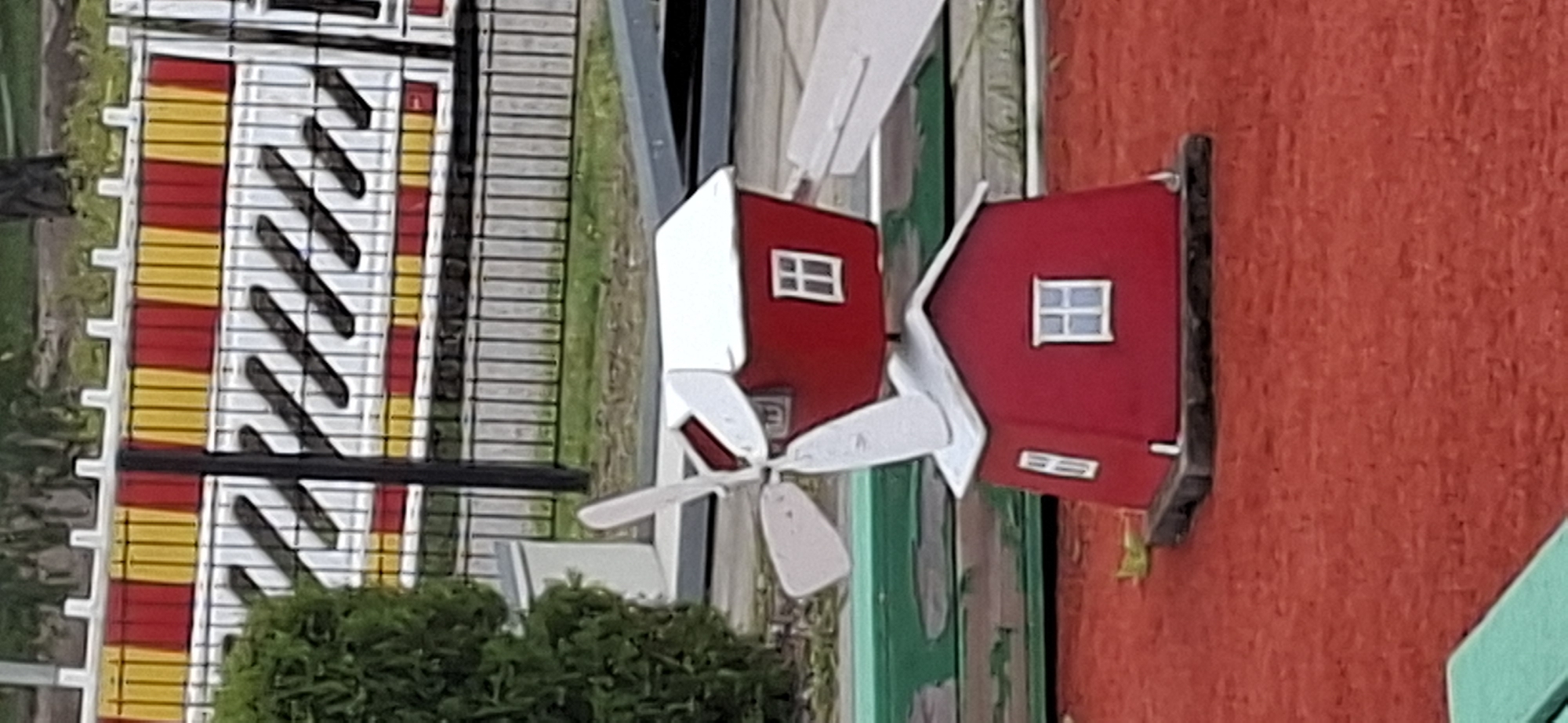 The image shows a miniature windmill decoration, likely a prop from a mini-golf course, situated on a red, sandy surface. The windmill has two tiers, both painted red with white roofs and white-framed windows. The top tier is angled, and attached to it are two white blades. The lower tier is a simple red house shape with a white roof and two windows. Behind the windmill, a black metal fence separates the foreground from a background with a white barrier decorated with alternating red and yellow stripes and black diagonal lines. Green grass and foliage are visible beyond the fence. The location is identified as a mini-golf course in Lahti, Finland. There are no people or animals present in the image.