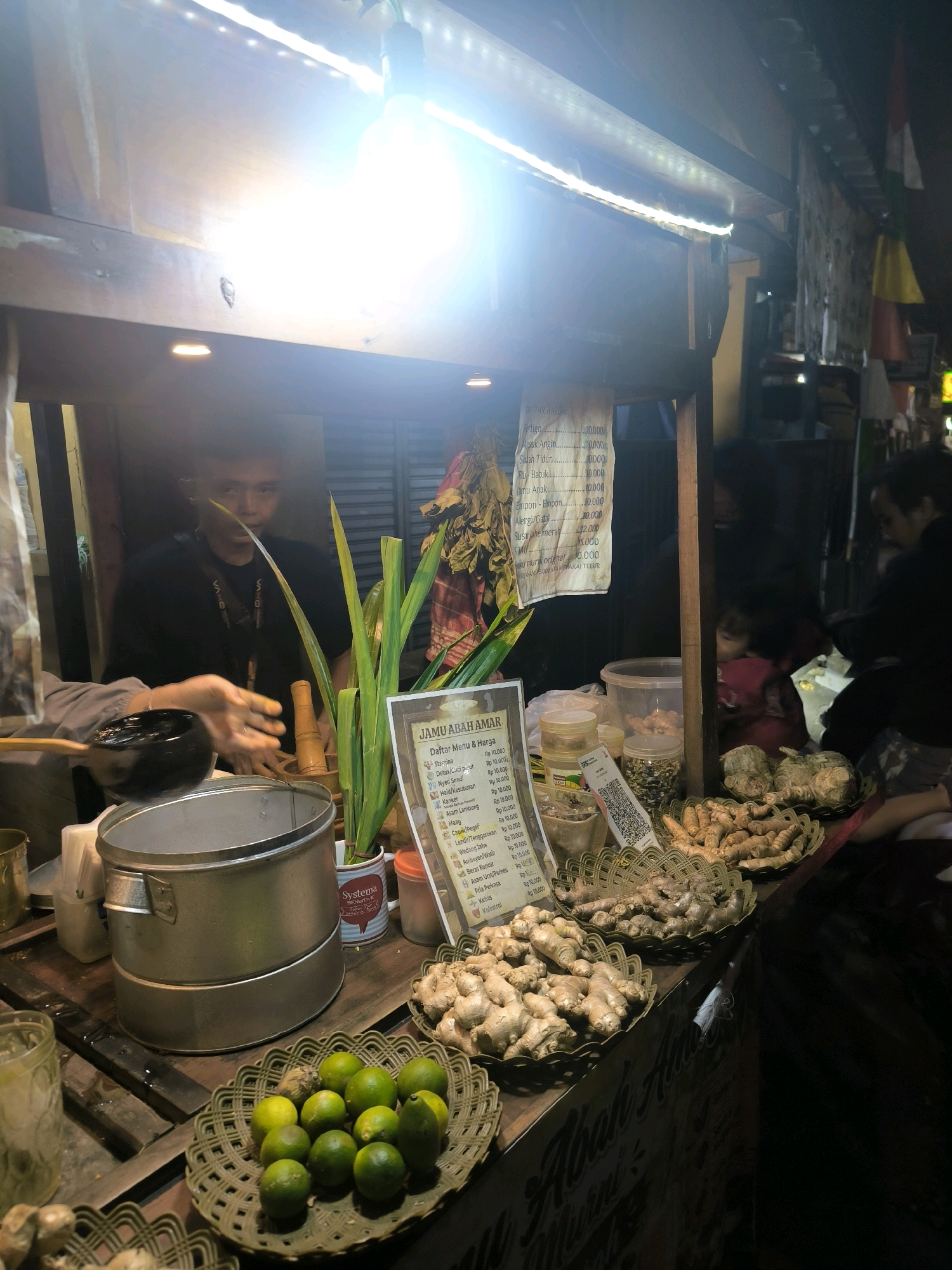 The image depicts a street-side stall in Bandung City, Indonesia, likely selling traditional herbal drinks, known as "jamu." In the foreground, a woven basket overflows with fresh limes, and another basket to the right holds a collection of ginger roots. Behind the limes, a large stainless steel pot is visible, suggesting a vessel for brewing or serving. To the right of the pot, a man in a black t-shirt is standing, with his hands near a wooden mortar and pestle, indicating he might be preparing ingredients. In front of him, a menu board is propped up, listing various herbal remedies with prices in Indonesian Rupiah. Further back, other customers, including a child, are dimly visible in the background, suggesting ongoing activity at the stall. The stall is illuminated by overhead lights and string lights, creating a warm and inviting atmosphere despite the dark surroundings of the night. The overall scene captures the essence of a local vendor providing traditional health remedies in an urban setting.