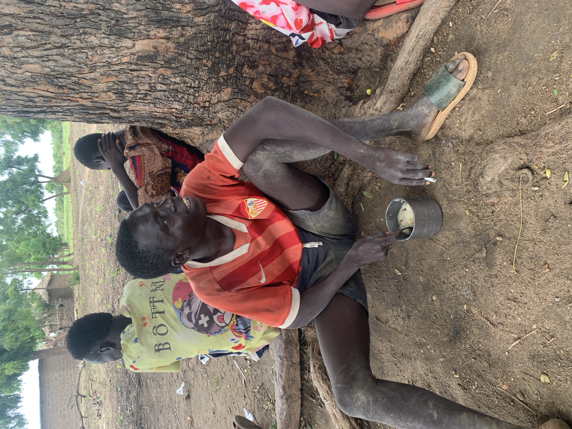 In Dull A, Nigeria, three individuals are seated outdoors near a large tree. The central figure, a dark-skinned male, wears an orange and red striped t-shirt and dark shorts. He is in a seated position with his legs crossed, holding a metal cup with a spoon. His right hand is near the cup, and his left hand holds a cigarette. His legs and arms appear to be dusty. To his left, another dark-skinned male wears a yellow t-shirt with the text "BOTTNI" printed on the back. To the right of the central figure, a third individual, whose face is not visible, wears a patterned fabric garment. The ground is composed of dirt and scattered debris, with tree roots visible. In the background, there are rudimentary structures that appear to be homes, and lush green trees. The lighting suggests it is daytime.