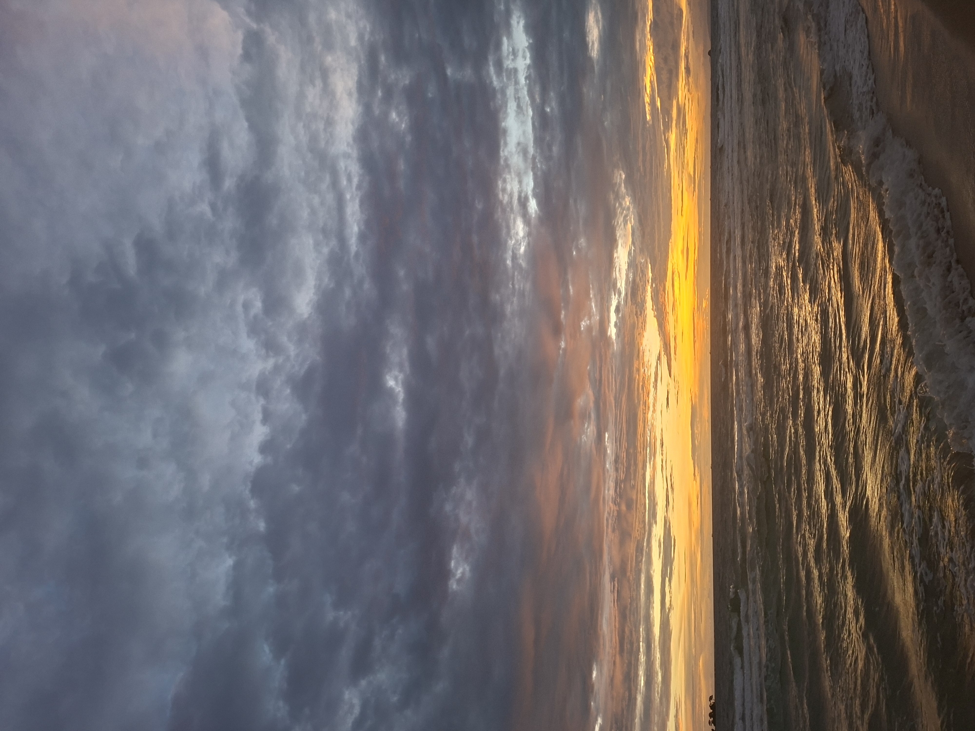 The image depicts a serene sunset scene over the ocean in Phú Quốc, Vietnam. The sky is a dramatic canvas of dark, brooding clouds gradually giving way to streaks of warm orange, yellow, and pink hues near the horizon. The ocean below reflects these vibrant colors, with gentle waves rolling towards a sandy shore. Small waves are visible crashing on the beach, creating a frothy white edge. In the distance, silhouettes of landmasses or islands can be seen on the horizon. The overall impression is one of tranquility and natural beauty during the twilight hours.