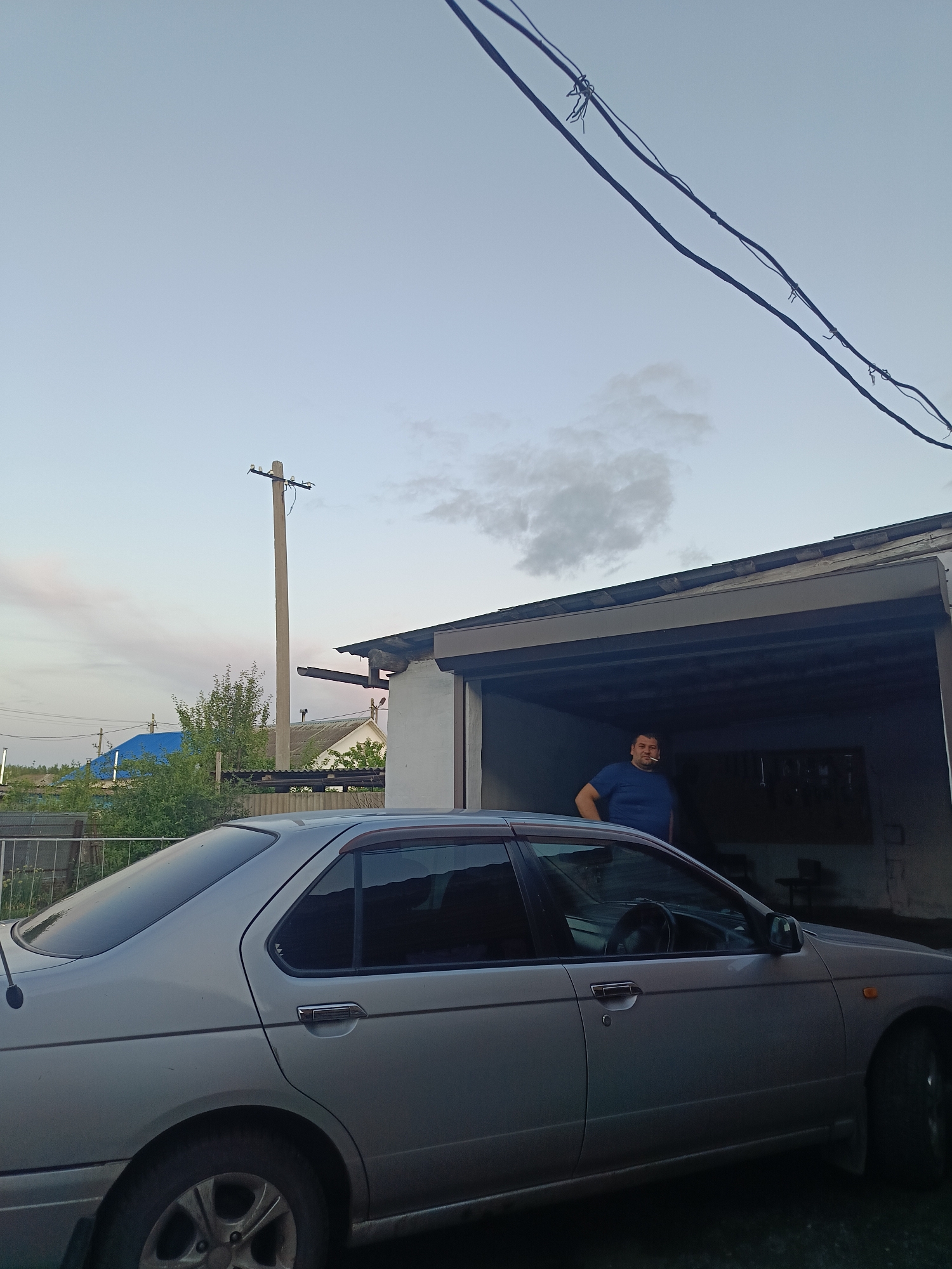 In the village of Khokhly, Russia, at dusk, a man stands in the open doorway of a garage, leaning against the frame. He wears a blue t-shirt. A silver sedan is parked in front of the garage, partially obscuring its entrance. Overhead, power lines crisscross the sky, and a utility pole stands to the left. In the background, houses with peaked roofs and a blue-tiled roof are visible amongst green trees. The sky is a pale blue with a few wispy clouds.