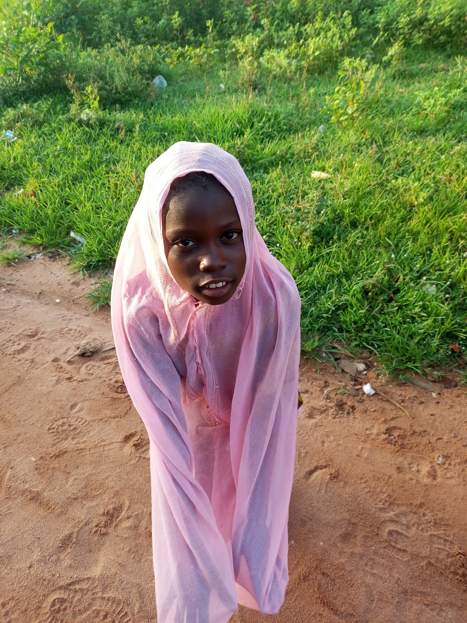 In a sandy area with sparse vegetation and a few pieces of litter, a young Black girl is pictured. She is wearing a light pink headscarf and a matching pink garment. Her eyes are dark and her mouth is slightly open, revealing her teeth. The lighting suggests it is daytime. The scene appears to be outdoors, possibly on the outskirts of a town or village.