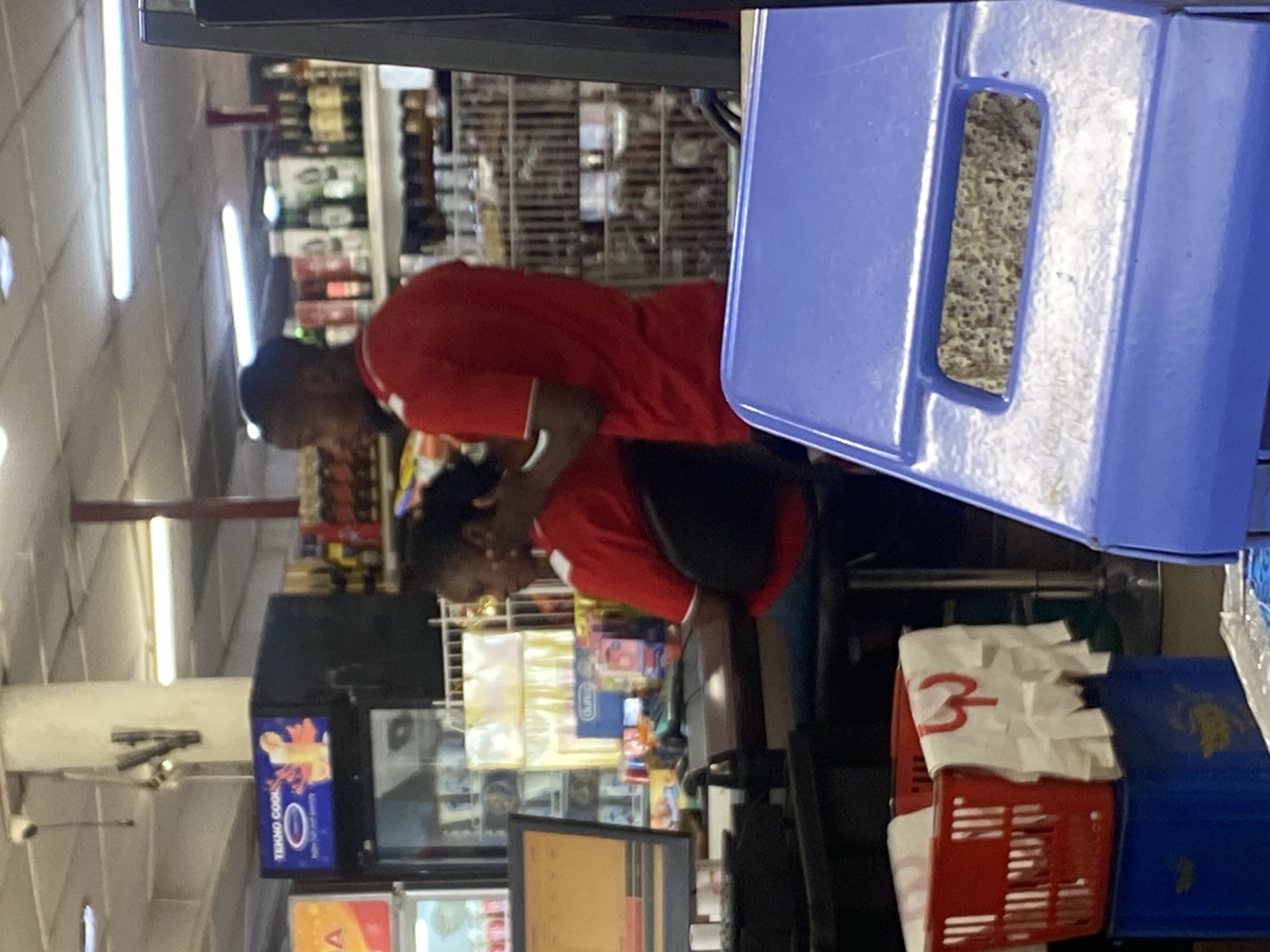 The image shows two people, a man and a woman, in what appears to be a store in Ikorodu, Nigeria. They are both wearing red shirts, and the man has his arm around the woman. The setting includes shelves stocked with various items, possibly drinks and snacks, and a blue plastic display unit. A red shopping basket is also visible. The lighting suggests it is daytime, likely indoors, as fluorescent lights are visible on the ceiling. The overall mood is difficult to ascertain from the image, but the close proximity of the individuals suggests a personal interaction. There is some visible text on packaging in the background and on a display in front of the couple, but it is largely illegible. One prominent piece of text on a white paper in the red basket reads "3-".