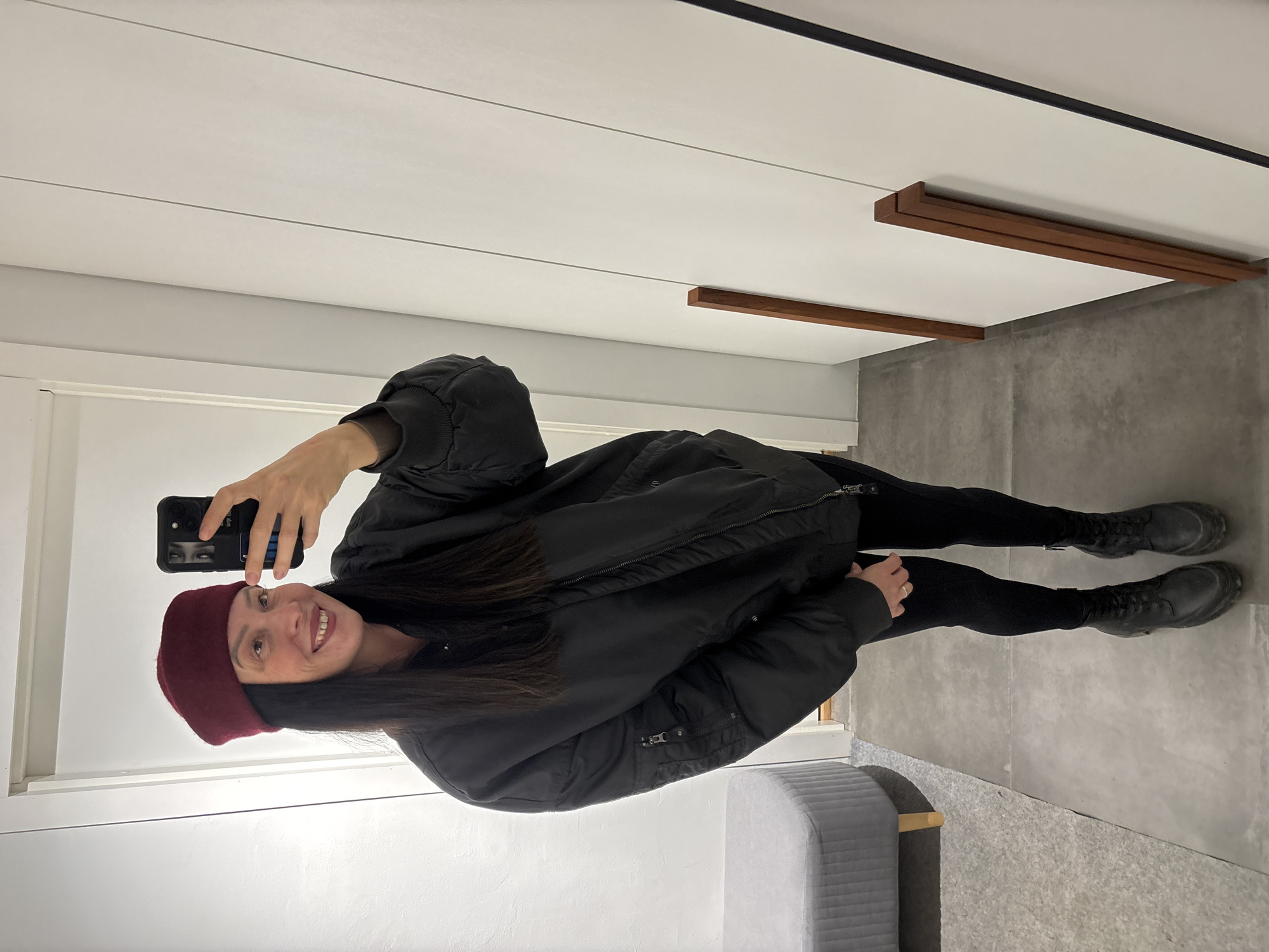 A woman stands in a room, taking a selfie with her smartphone. She wears a burgundy beret, a black puffy jacket, black leggings, and black boots. The room has a grey concrete floor and white walls with wooden accents. A grey ottoman sits in the corner. The location is possibly in the city of Chaikovsky, Russia.