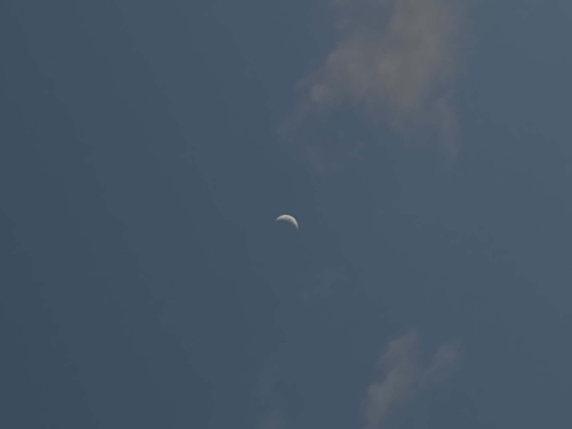 The image is a vertical, medium shot of the sky at dusk, with a sliver of the moon visible. The sky is a deep, clear blue, with a few wispy clouds scattered across the frame. The moon, a thin crescent, is located slightly left of the center of the image, glowing faintly against the darkening sky. The overall atmosphere is serene and peaceful. The location is unknown.