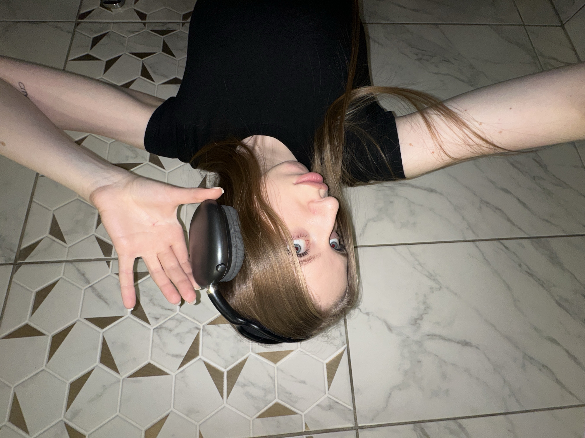 A young woman lies on a tile floor. The tiles are patterned with white hexagons containing golden triangles, and gray marble tiles surround the patterned area. She wears a black top, and her blonde hair is spread around her head. Black headphones rest on her ear, and one hand is held up, fingers outstretched. A tattoo is visible on one of her arms. The image is inverted. The location is unknown.