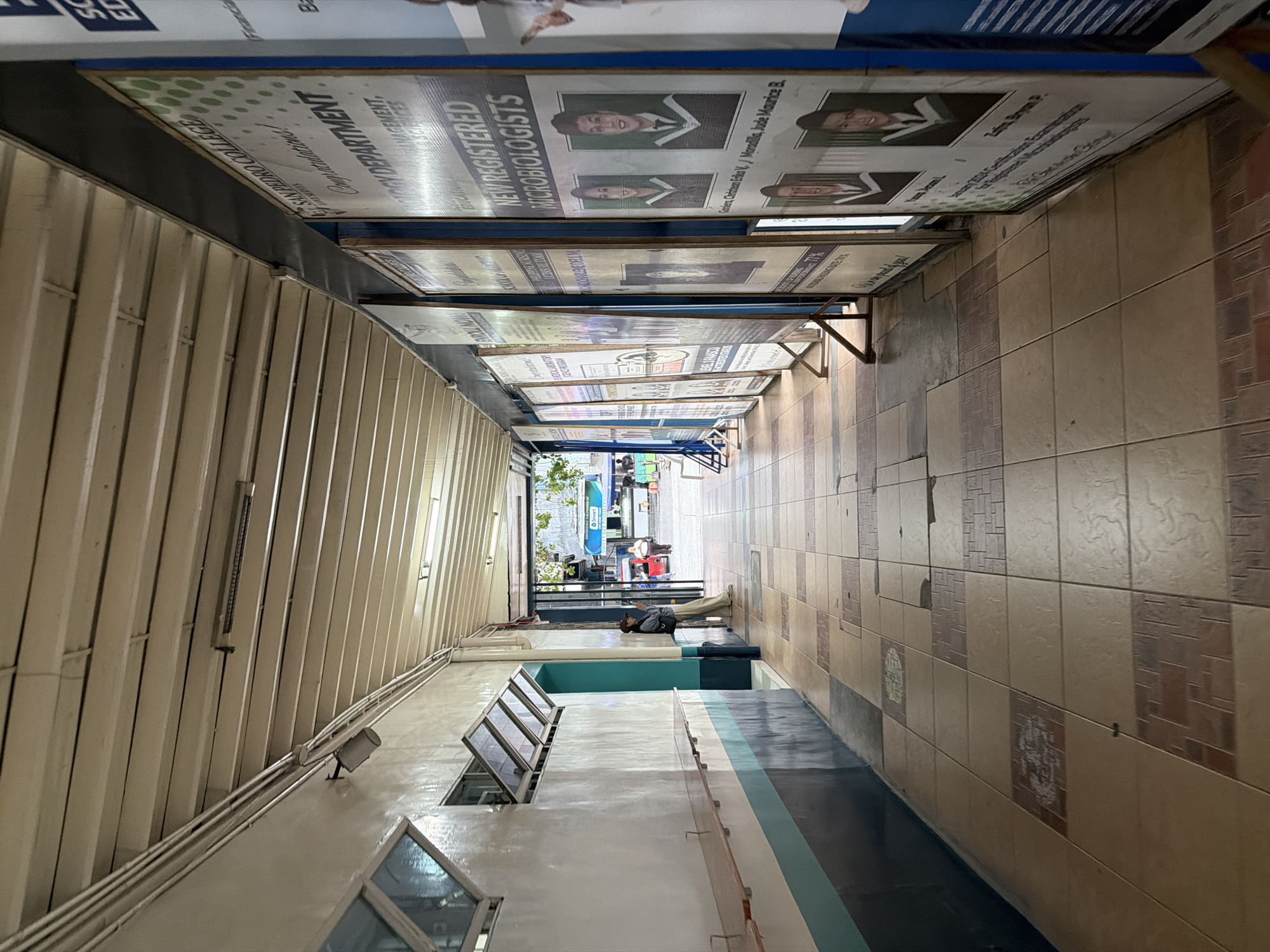 The image depicts an outdoor walkway at San Pedro College in Davao City, Philippines. The walkway is covered by a beige, ribbed ceiling, with pipes and light fixtures visible. On the left, a white wall is painted with horizontal stripes of teal and dark gray. Two windows with open angled glass panels are present on this wall. A wooden railing runs along the wall at waist height. The floor is tiled with a pattern of beige, brown, and reddish-brown squares.
On the right side of the walkway, a series of large posters are displayed vertically, supported by metal stands. These posters appear to be celebratory announcements, possibly from the college, featuring photographs of students in academic regalia and text. One prominent poster announces "NEW REGISTERED MICROBIOLOGISTS" and displays the names and faces of several individuals.
In the middle ground of the walkway, a person wearing a baseball cap, a light-colored jacket, and loose-fitting pants is leaning against the wall, looking down at a mobile phone. They have a black bag slung over their shoulder. Further down the walkway, the scene opens to an outdoor area with trees, more posters, and people in the distance, including a red vehicle and a bus with a "Smart" advertisement. The overall impression is that of a functional, somewhat utilitarian campus hallway used for displaying academic achievements.