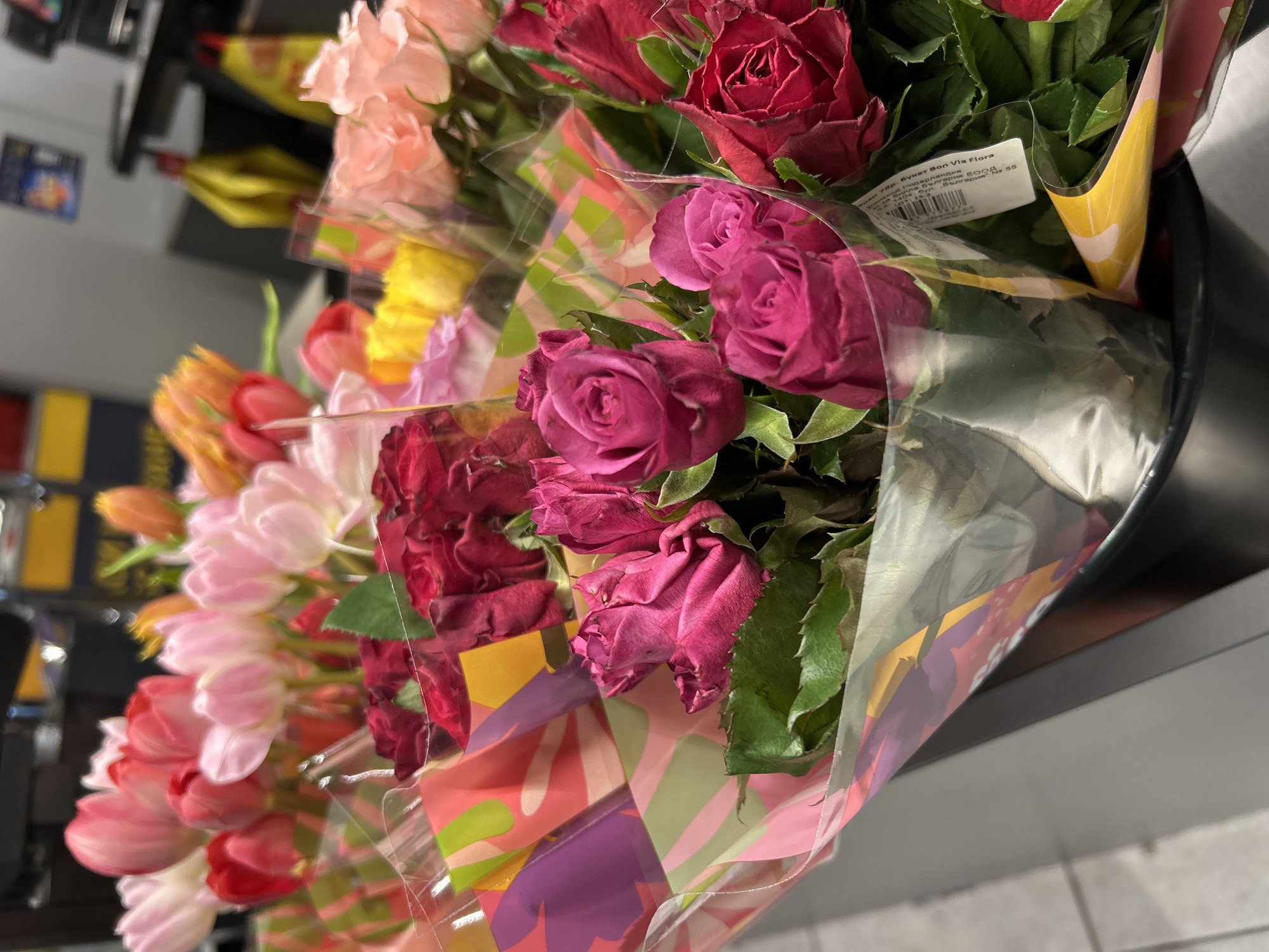 The image shows a close-up view of multiple bouquets of flowers arranged in a container, likely for sale in a store. The bouquets contain a variety of flowers, including roses (in shades of red and pink) and tulips (in shades of pink, orange, and red). The flowers are individually wrapped in clear plastic with patterned paper underneath. A label is visible on one of the bouquets. The setting appears to be indoors. Based on the visible text on the labels, the bouquet is a "Букет Bon Via Flora", making it likely to be located somewhere in Bulgaria. Given the user's prompt, the image is likely taken in Plovdiv.