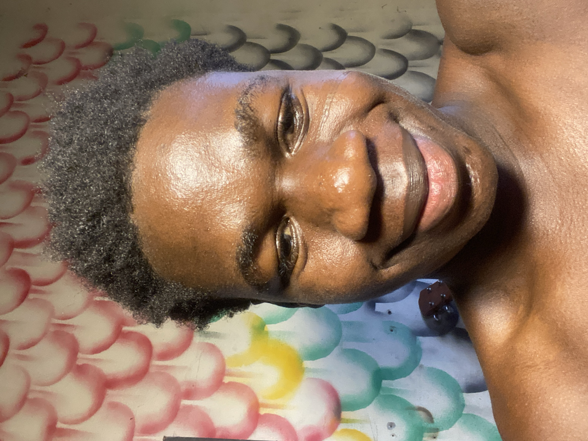 A close-up, slightly angled, and brightly lit photograph features a person with dark brown skin. The person has short, dark hair. The person is smiling slightly. The background is a surface painted with multi-colored circles. Part of the person's shoulder is visible. A blurry object resembling a small container is also present in the background. It is not possible to establish the location in Mararaba, Nigeria.