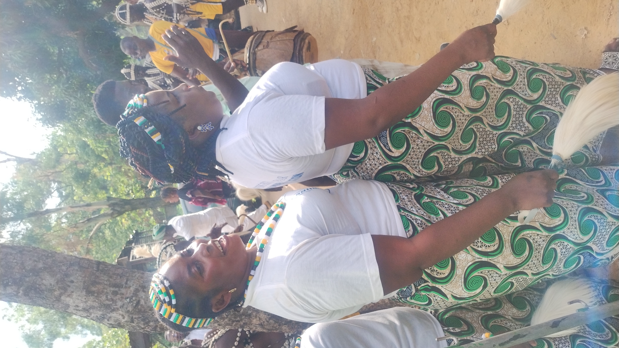 This image depicts an outdoor scene, likely in Jos, Nigeria, with several individuals, predominantly women, participating in what appears to be a cultural event or performance. Two women in the foreground are adorned with vibrant, beaded headbands and necklaces, and are wearing white t-shirts paired with elaborately patterned skirts featuring swirling green and cream designs. They are holding white, whisk-like objects, possibly for dancing or ceremonial purposes.
In the background, other people are visible, some wearing traditional attire. One man in a yellow shirt is standing next to a drum, suggesting a musical component to the event. The environment is characterized by lush trees and a rustic, earthy ground. The lighting suggests it is daytime, possibly with the sun casting shadows and creating a bright atmosphere. The overall impression is of a lively gathering, rich in cultural expression.