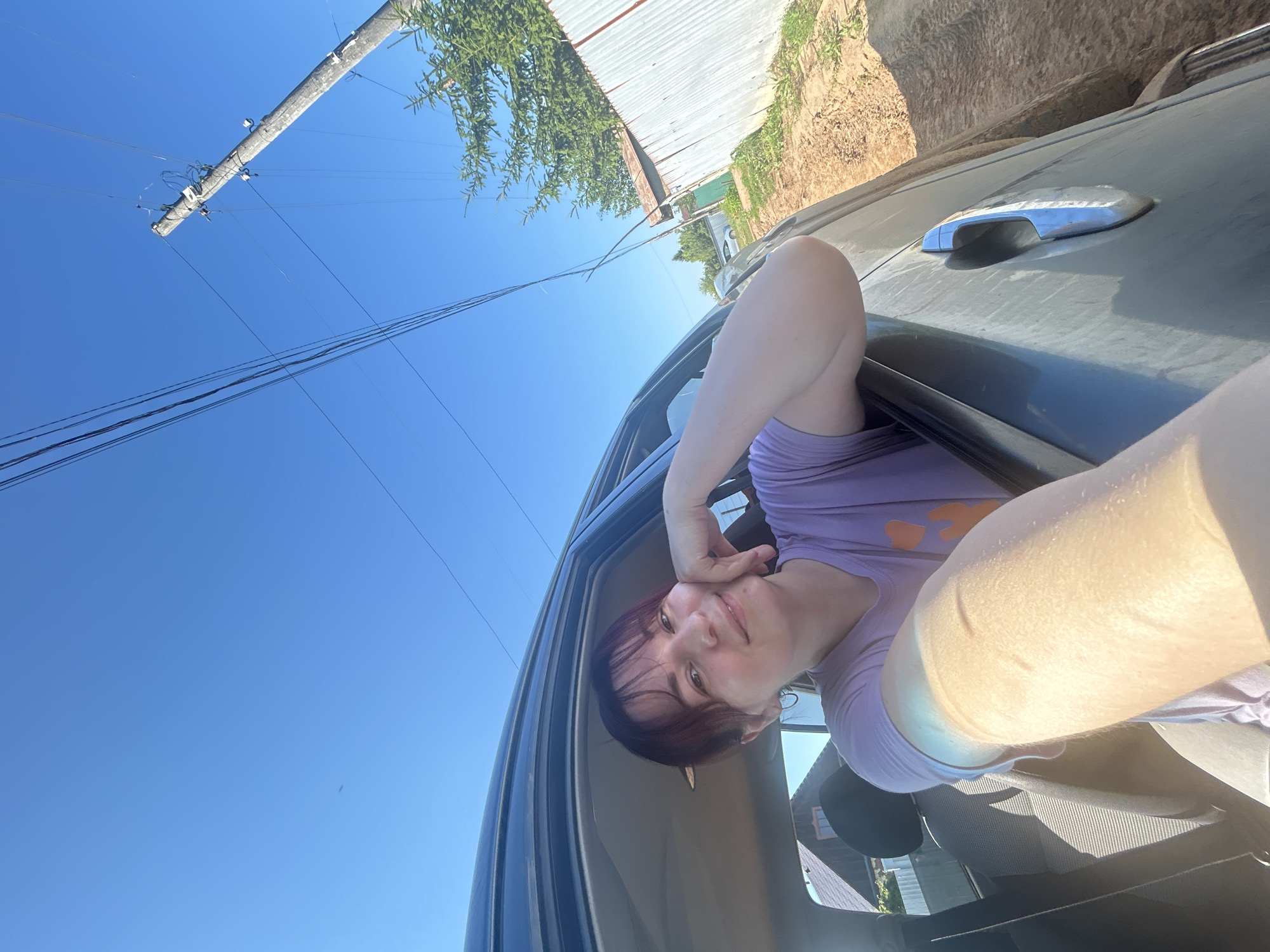 A young woman with dark hair and bangs is leaning out of a car window, smiling at the camera. She is wearing a light purple t-shirt. The car is on a dirt road, with a corrugated metal fence and some trees in the background. A utility pole with wires is visible against a clear blue sky. The scene appears to be in a rural area, possibly in the city of Fokino, Russia, given the context. The sunlight suggests it is daytime.
