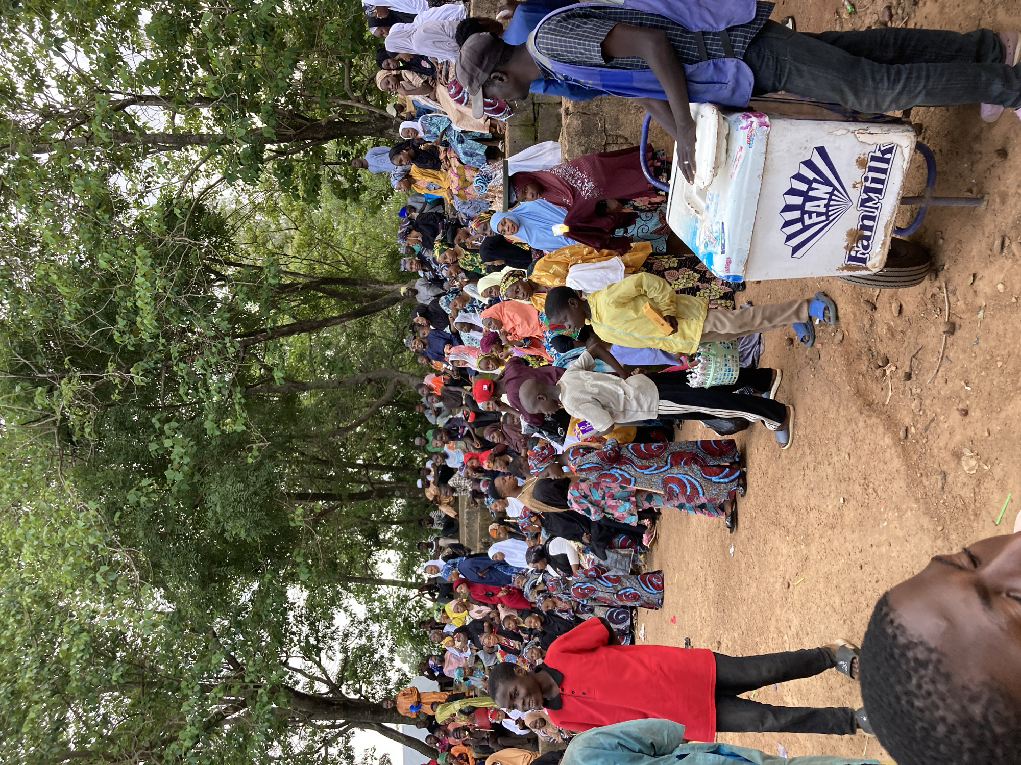 The image captures a gathering of people in an outdoor setting, likely a community event or a market, with a backdrop of lush trees. The crowd, composed of adults and children, is predominantly dressed in colorful traditional attire, including headscarves and patterned fabrics. Some individuals are seated on tiered, natural rock formations, while others stand. In the foreground, a young boy in a red shirt is prominently positioned, looking to his left with a slight smile. To his right, another young boy in a yellow jacket holds a small yellow object. A man wearing a blue vest and a dark cap stands near a white cooler with the "Fan Milk" logo, appearing to be selling beverages. The ground is covered in a sandy, reddish-brown dirt. The overall atmosphere suggests a lively, communal event taking place in Jos, Nigeria, a city known for its vibrant cultural activities and historical indigenous settlements.