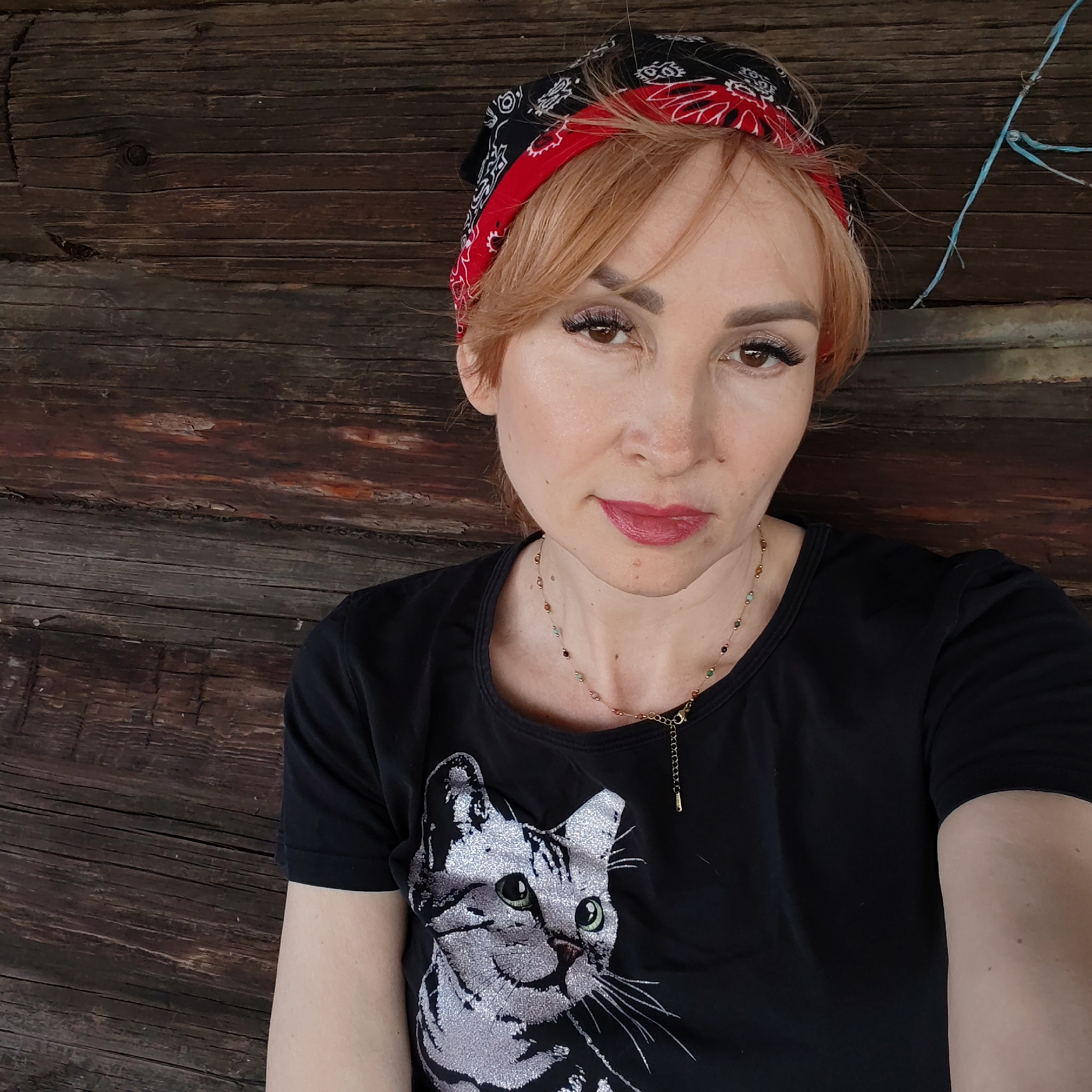 A woman stands close to the camera, centered in the frame, positioned against a rough, dark wooden wall. She is wearing a black T-shirt with a large, sparkly silver cat graphic on the front. Her hair is reddish-blonde, and she wears a black and red patterned bandana. She has a delicate necklace with colorful beads.
In the background, to the right of the woman, a fragment of blue string or rope is visible against the wood. This photograph seems to be taken outdoors, with the rough wooden planks acting as a backdrop. The woman's posture and expression suggest a casual selfie. It is not possible to confirm the location of Chaikovsky, Russia based on the visual data provided.