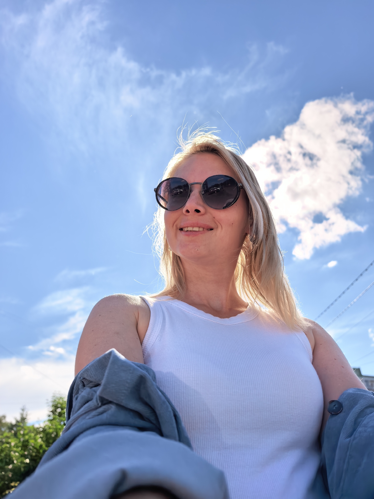 The image depicts an adult female captured from a low-angle perspective in an outdoor setting, against a clear blue sky with scattered white clouds. The subject has blonde hair, back-lit by the sun, and wears dark, round-framed sunglasses. She is smiling faintly, with her mouth slightly open, revealing teeth, and looking towards her left (viewer's right). Her attire consists of a white ribbed tank top. A blue garment, which appears to be a jacket or shirt, is draped over her right arm (viewer's left), displaying a dark button on its sleeve. In the background, green foliage is visible on the lower left, and multiple thin, dark lines, consistent with power lines or utility cables, traverse the upper right sky. The scene is illuminated by bright natural sunlight. The location is specified as the city of Sterlitamak, Russia; however, the visual information present in the image does not provide explicit identifiers to definitively confirm this specific geographical location.