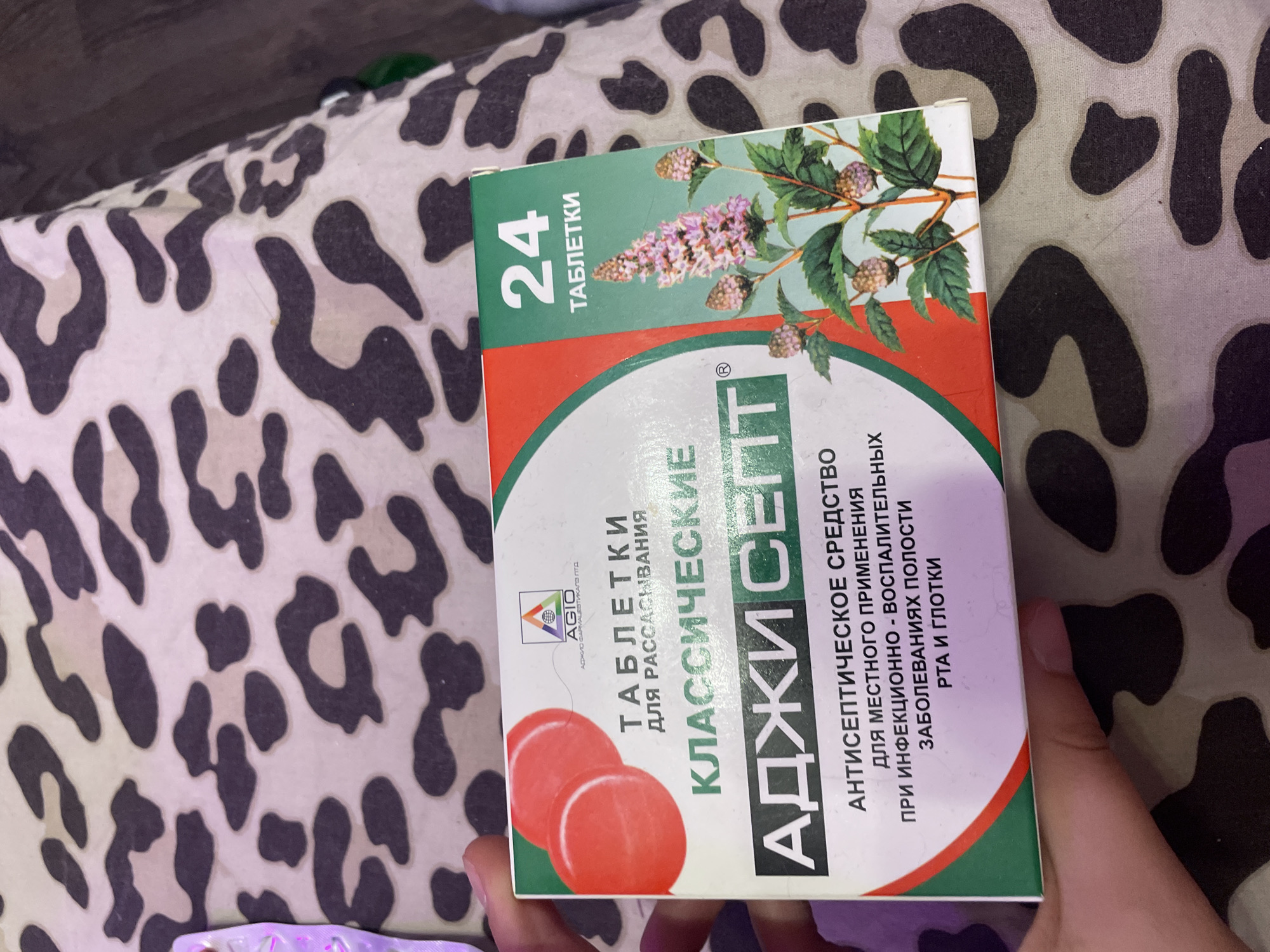 A person's hand is holding a box of medication. The box is predominantly white with green and red accents. The front of the box features the brand name "АДЖИ СЕПТ" in large, bold lettering, with "КЛАССИЧЕСКИЕ" above it. Below the brand name, it states "АНТИСЕПТИЧЕСКОЕ СРЕДСТВО ДЛЯ МЕСТНОГО ПРИМЕНЕНИЯ ПРИ ИНФЕКЦИОННО - ВОСПАЛИТЕЛЬНЫХ ЗАБОЛЕВАНИЯХ ПОЛОСТИ РТА И ГЛОТКИ". There is also an image of a flowering plant on the right side of the box, and a logo with "AGIO" and Russian text. On the upper right corner, it indicates "24 ТАБЛЕТКИ". The box is placed on a surface with a leopard print pattern. The image is a close-up shot, with the background blurred. The specific location within the city of Vaganovo, Russia is not discernible from the image.