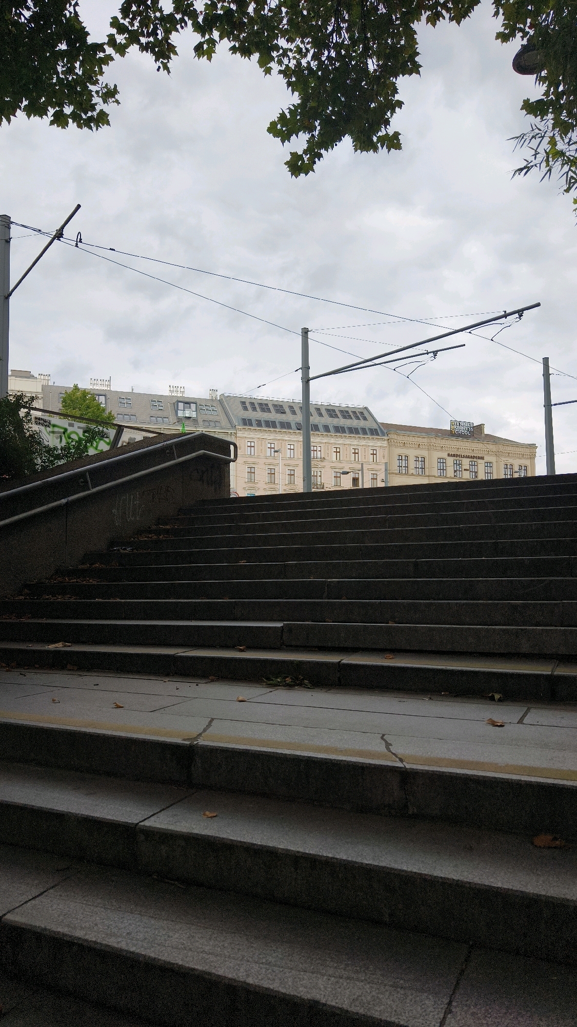 The image shows a view from the bottom of a wide outdoor staircase looking upwards towards buildings and a cloudy sky. The staircase is made of dark stone with visible joints and some scattered autumn leaves on the steps. A metal handrail runs along the left side of the stairs. Above the staircase, there are several multi-story buildings with traditional architecture, characterized by numerous windows and pitched roofs. Overhead, there are overhead power lines supported by metal poles, likely for a tram system. The overall atmosphere is overcast and perhaps a bit damp, consistent with autumn weather. The buildings in the background are light-colored, with some details suggesting the location is in Vienna, Austria, as indicated by signage visible on one of the buildings that reads "VIENNA BUSINESS SCHOOL" and "HANDELSKAKADEMIE".