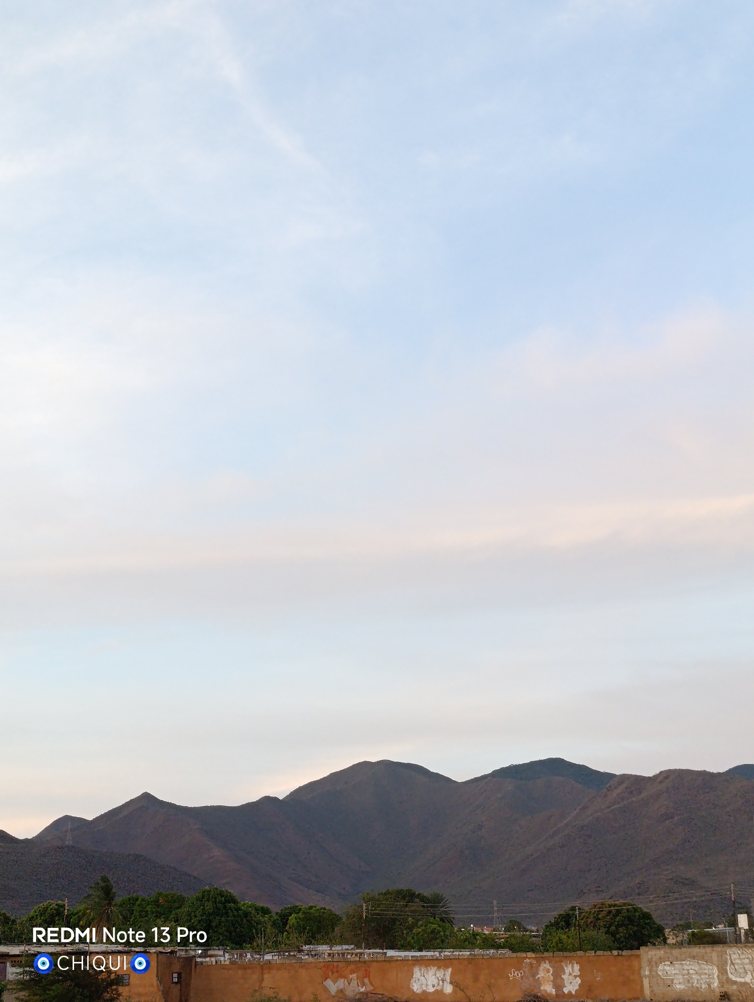 The image depicts a mountainous landscape in the city of Cotoperíz, Venezuela. The mountains are brown and green, and they are silhouetted against a light blue sky. In the foreground, there is a low wall with some trees behind it. The sky is pale blue. The image was captured by a REDMI Note 13 Pro, identified as taken by "CHIQUI."