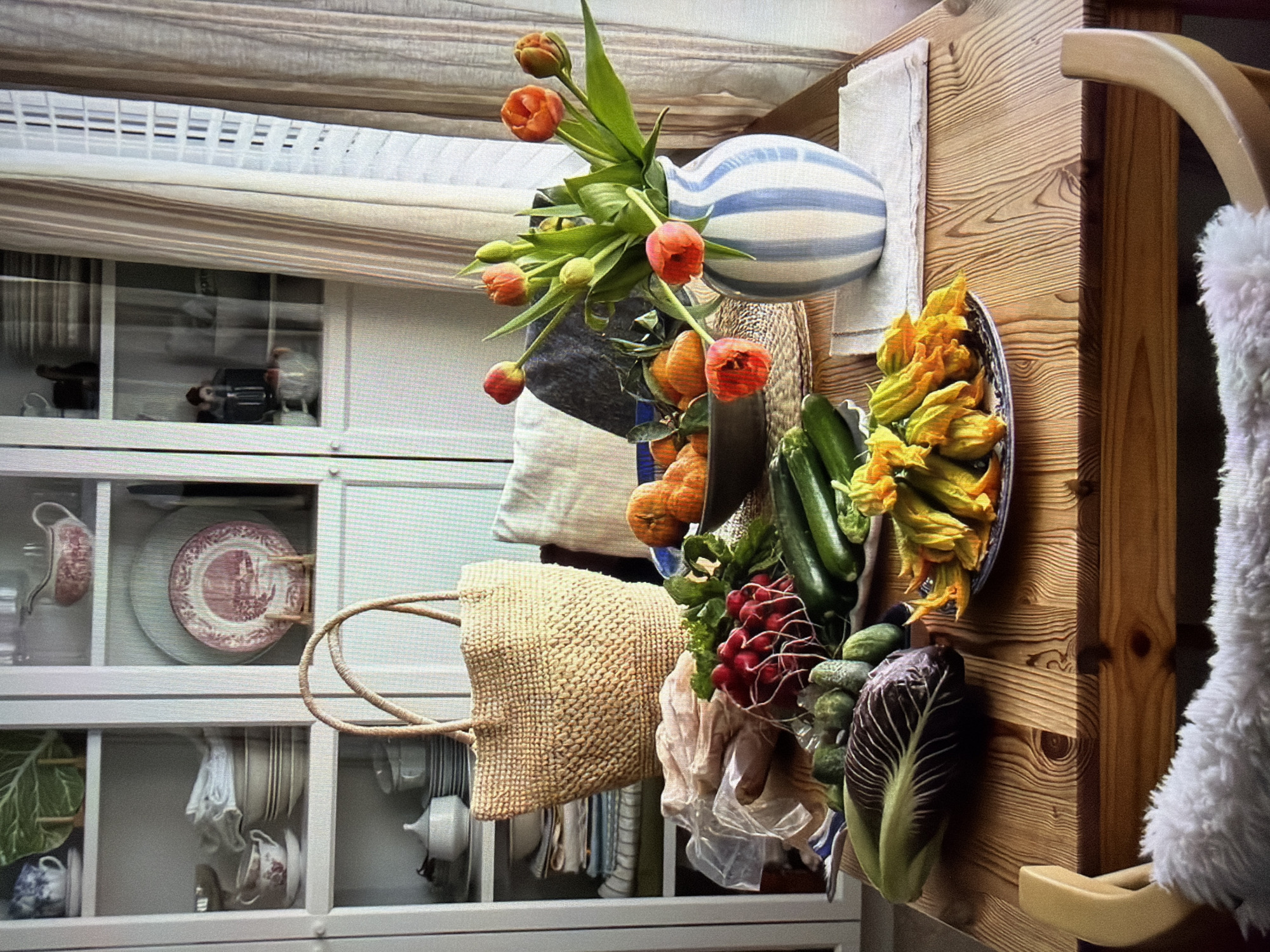 This image, taken in Ros Na Rí, Ireland, captures a vibrant indoor scene, oriented vertically. A rustic wooden table or counter dominates the right side of the frame, featuring a strong wood grain pattern. Attached to the table is a light wooden hook holding a soft, fuzzy white object, possibly a garment or blanket.
The table is laden with an assortment of fresh produce and decorative items. A blue and white striped ceramic vase holds a bouquet of bright orange-red tulips, placed on a light-colored linen cloth. Beside the vase, a large oval dish, patterned in blue and white, overflows with delicate bright yellow zucchini blossoms. A bunch of dark green zucchinis, red radishes with their greens, and small gherkins are artfully arranged nearby. A distinct head of radicchio, with its deep purple and white leaves, rests at the forefront of the produce. Oranges or small pumpkins are partially visible behind the vase. A woven straw tote bag hangs over the edge of the table, and a transparent plastic bag containing unidentifiable items is nestled amongst the produce.
In the background, to the left, a white wooden cabinet or shelving unit with glass doors displays various dishware. Visible items include plates with a red/pink pastoral pattern, plain plates, teacups, saucers, and bowls, some matching the patterned plates, others in white or blue and white designs. Green decorative leaves are also placed within the shelves. Above the cabinet, a window with reflective glass shows indistinct objects or reflections, and horizontal blinds or shades are visible at the top.
The scene suggests a domestic setting, possibly a kitchen or dining area, showcasing fresh ingredients, implying a recent market visit, a harvest, or preparation for a meal. The arrangement creates a warm, inviting, and abundant atmosphere, characteristic of a cozy home environment. No people are visible, and no discernible text appears in the image. The time of day is not explicit, but natural light is present.