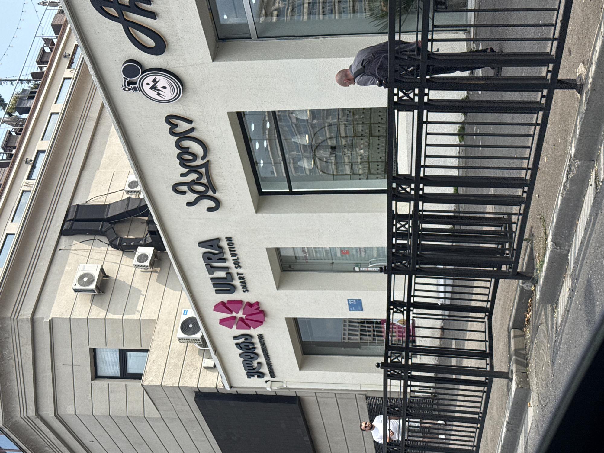 This image captures a storefront on a street in Tbilisi, Georgia. The building is modern with a light-colored facade. Above the storefront, there are prominent signs with text and graphics. One sign reads "ULTRA SMART SOLUTION" with a stylized pink logo resembling a flower or abstract shape. To the right of this, another sign features Georgian script and a circular logo depicting mountains within a compass-like design. To the far right, part of another business's sign is visible with bold, cursive lettering.
Below the signs, there are large display windows. Through the windows, interior lighting and some decorative elements are visible, including what appears to be a wrought-iron structure or display case.
In front of the storefront, a black metal railing runs along the sidewalk. On the sidewalk to the left, a man wearing a white t-shirt and shorts stands looking towards the camera. To the right, further down the sidewalk, a bald man wearing a grey shirt and dark pants with a backpack is leaning on the railing, looking towards the building.
The street in front of the railing is paved, with a concrete curb and what appears to be a manhole cover. The overall scene suggests a commercial district within the city of Tbilisi.