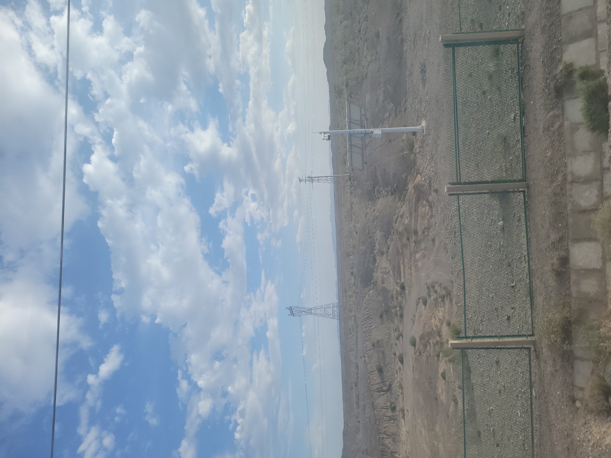 The image depicts a wide outdoor landscape with a focus on power lines and transmission towers against a backdrop of a vast, hazy sky filled with cumulus clouds. The scene is oriented vertically, suggesting it may have been taken from a high vantage point or with the camera tilted.
In the foreground, a section of a metal fence with green mesh and wooden framing runs across the ground, which is covered in gravel and sparse vegetation. Adjacent to the fence are paved areas or concrete structures.
Further into the scene, a large, lattice-style transmission tower stands prominently, supporting multiple power lines that stretch across the frame. In the distance, a more modern, single-pole utility structure with attached equipment is visible. The terrain is arid and mountainous, with the hills appearing barren and rocky, extending towards the horizon where they merge with the hazy sky.
The time of day appears to be daytime, given the bright illumination and the presence of distinct clouds. The weather seems to be fair but hazy, obscuring distant details. The location appears to be a somewhat remote, possibly industrial or infrastructure-heavy area within a desert or mountainous region. No people are visible in the image. The overall impression is one of infrastructure in a natural, rugged environment.