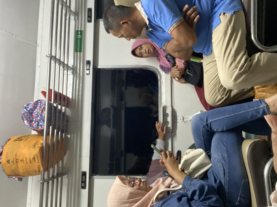 The scene depicts the interior of a passenger train carriage, specifically within the city of Banjarwungu, Indonesia. Four individuals are visible within the frame. On the left, a female wearing a light pink hijab, eyeglasses, a blue denim shirt, and blue jeans sits, looking down at a green-cased smartphone held in her right hand. A white handbag rests on her lap. Her bare feet are partially visible in the lower left. Behind her, a child or young person's hand rests on the window ledge of the train. To the right, an older female, also wearing a pink hijab and a dark purple top, is seated, looking forward towards the male beside her. She holds a black bag on her lap. Next to her, an older male wearing a blue polo shirt with striped trim and light brown trousers is seated with his arms crossed, facing left. The train interior is white, featuring a dark tinted window. An overhead metal luggage rack contains an orange bag, a blue and white patterned bag, and a red object. Seat numbers or section indicators "8DE," "7DE," and "6DE" are visible above the window, along with a green directional sign. The individuals appear to be passengers engaged in travel, with one focused on a device and the others observing or interacting.