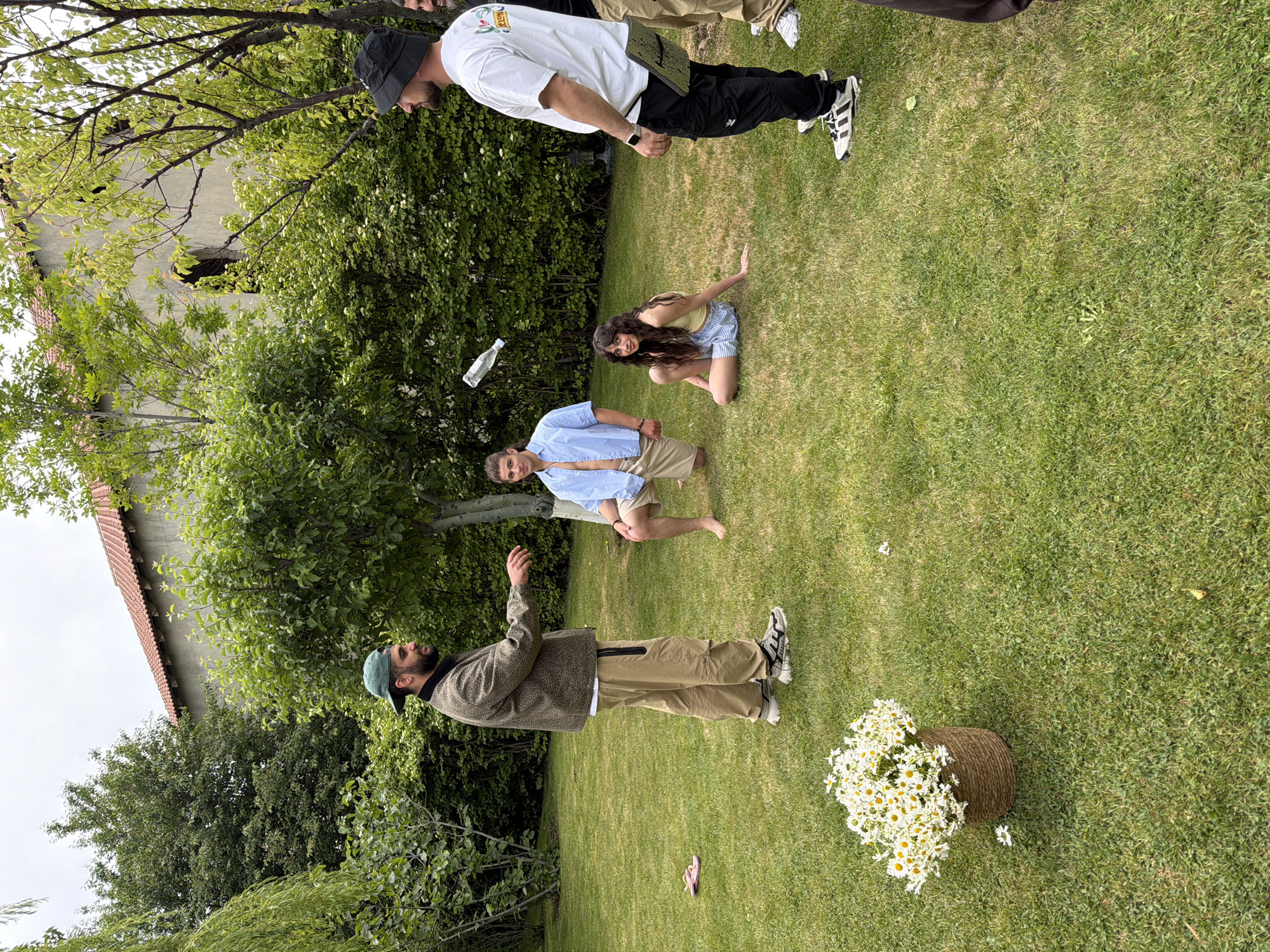 The scene is set in a grassy yard in Yerevan, Armenia, where four individuals are present. One man, wearing a green cap and a khaki-colored jacket and pants, is standing with his arm raised as if gesturing or throwing something. A plastic bottle is suspended in mid-air between him and a man in a light blue shirt and khaki shorts, who is also standing. On the ground, a woman with long dark hair is kneeling, looking toward the camera. Another man stands above her, wearing a white t-shirt, black pants, and a black cap. A wicker basket filled with daisies sits in the bottom right corner of the scene. In the background, there is a building with an orange tile roof and trees.