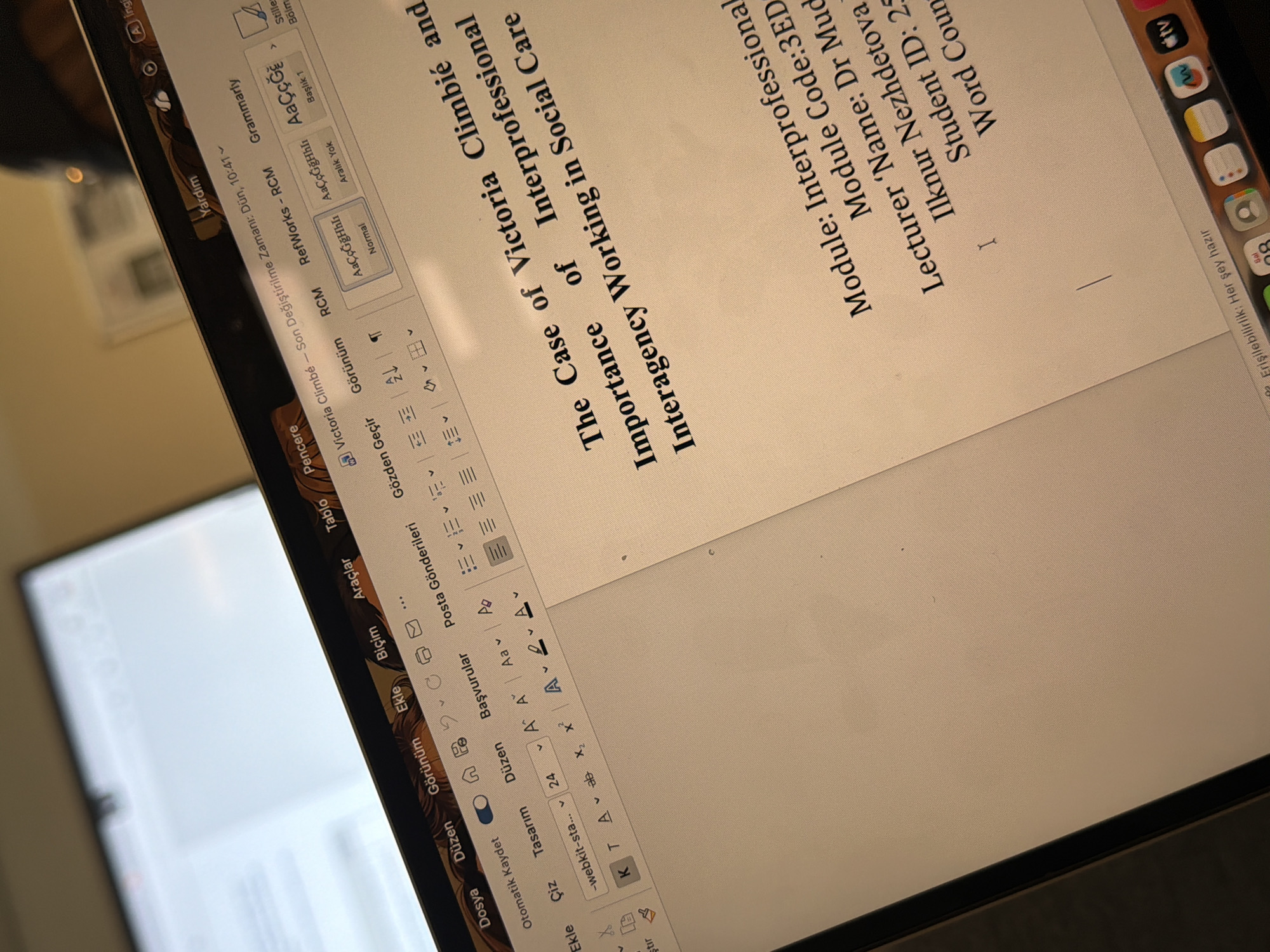 The image displays a close-up, angled shot of a laptop screen featuring a word processing document, likely an academic paper, in an indoor setting. The screen is captured from an oblique, high-angle perspective, with reflections on the left side. In the out-of-focus background, another screen or monitor with a white display is visible.
The document on the laptop screen is titled "The Case of Victoria Climbie and Importance of Interprofessional Interagency Working in Social Care". Visible academic details include "Module: Interprofessional", "Module Code:3ED", "Lecturer Name: Dr Mud Ilknur Nezhdetova", "Student ID: 25", and "Word Coun" (presumably Word Count). The document's last modification is noted as "Dün, 10:41" (Yesterday, 10:41).
The word processing software's interface, resembling Microsoft Word, is in Turkish, showing menus like "Dosya" (File), "Düzen" (Edit), "Görünüm" (View), "Ekle" (Insert), "Biçim" (Format), "Araçlar" (Tools), "Gözden Geçir" (Review), "RCM", and "Grammarly". Formatting options such as "Normal", "Aralık Yok" (No Spacing), and "Başlık 1" (Heading 1) are also visible, with a font size set to "24".
Part of the macOS dock is visible on the right side of the laptop bezel, displaying icons for applications including TV, Messages, Safari, Maps, Calendar, and App Store. The macOS status bar in the upper right shows "Sal" (Tuesday) and the message "Erişilebilirlik: Her şey hazır" (Accessibility: Everything is ready). No individuals are visible, but the scene suggests active use of the laptop for study or work. The environment implies a study or office space, with the provided context indicating a location in Sunderland, United Kingdom, though no specific visual elements within the image confirm this.