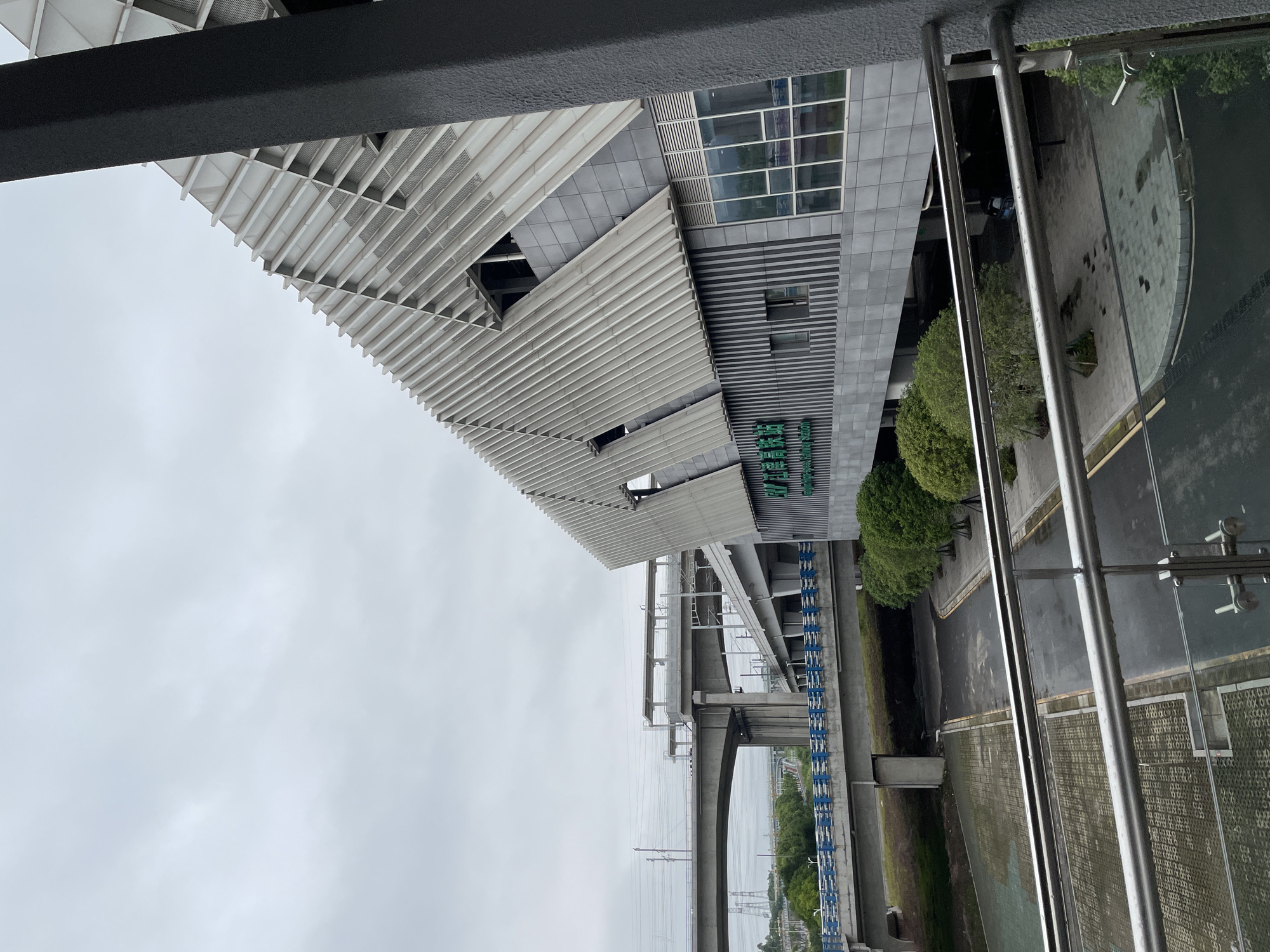 The image depicts a partially obstructed view of what appears to be a modern transportation hub, identified by the sign in Chinese as a Jiangjin station, presumably in Jiangjin, China. The building features a distinctive facade with a series of horizontal louvers above a gray façade section with windows.
A elevated bridge structure with blue and white barriers and power lines can be seen towards the left of the station. A gray sidewalk and road are visible near the station.
The image is taken from an elevated position, possibly a building or a bridge, with foreground structures obstructing the view.
The sky appears overcast.