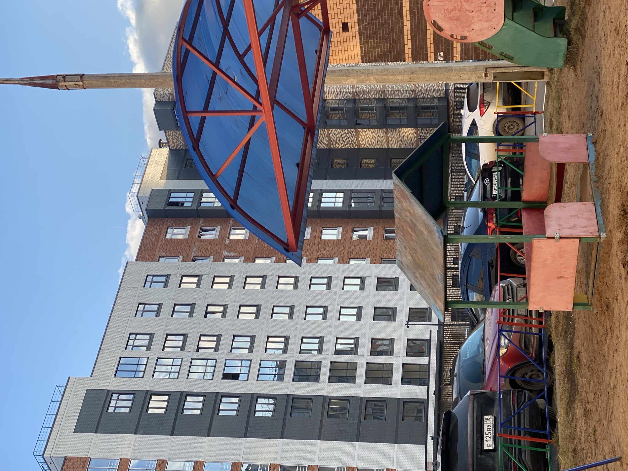 The image shows a playground in Izhevsk, Russia, located near a multi-story residential building with a brick facade in varying shades of gray, white, and brown. The playground equipment includes a blue-covered structure with a red frame, a children's playhouse made of wood, and a slide with a green platform and red accents. Several parked cars are visible behind the playground equipment. The ground appears to be covered in sand. The sky is blue with some clouds. A tall concrete pole extends upward into the sky. A license plate is visible on one of the cars.