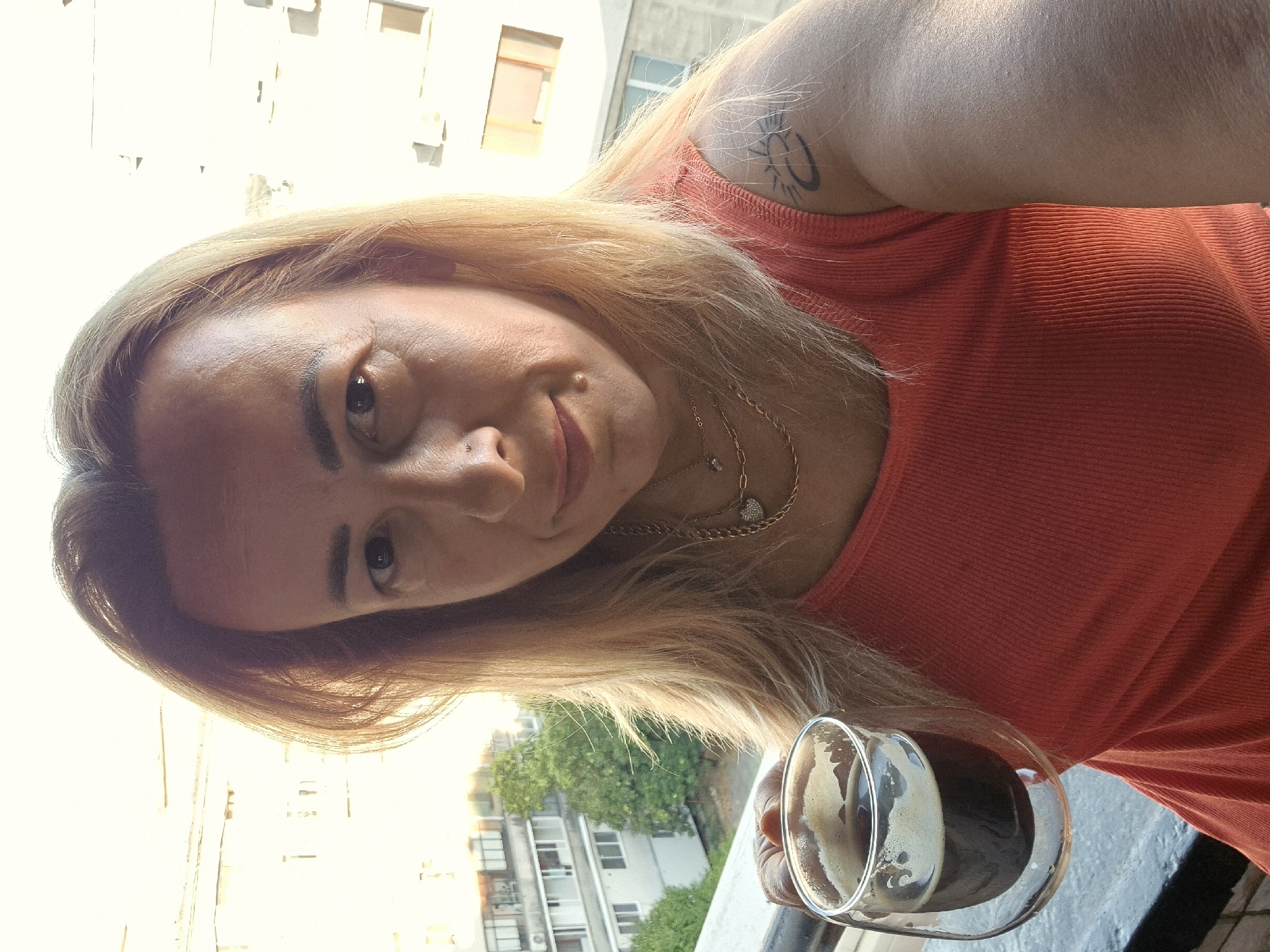 A medium shot captures a woman with blonde hair and a pink tank top holding a glass of dark liquid, possibly coffee, while standing on a balcony. The woman's face is angled towards the camera, and she has a faint smile. She is wearing multiple necklaces. A tattoo is visible on her right shoulder. In the background, apartment buildings and trees are visible under a bright sky, suggesting an urban outdoor setting. The light indicates it is daytime.